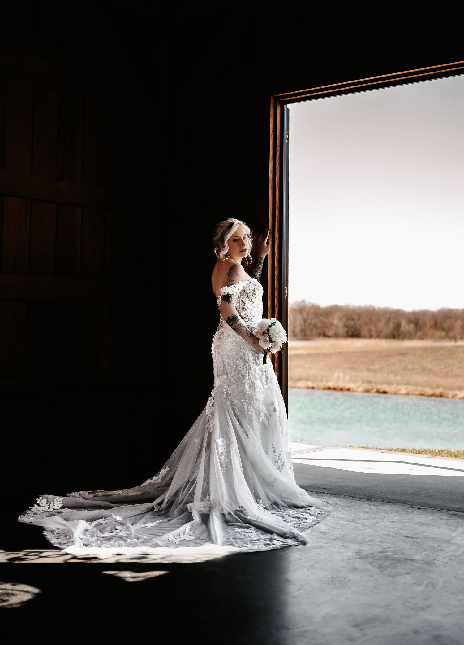 Bride in elegant lace mermaid wedding gown with long train stands in barn doorway, holding bouquet, overlooking scenic countryside.