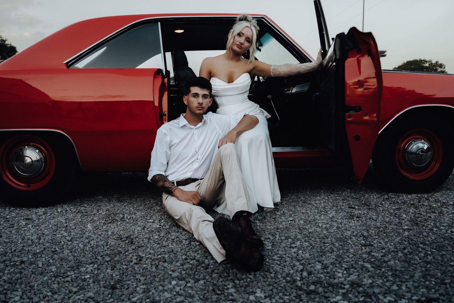 Couple in wedding attire posing by a classic red car, bride in white dress seated on groom in white suit.