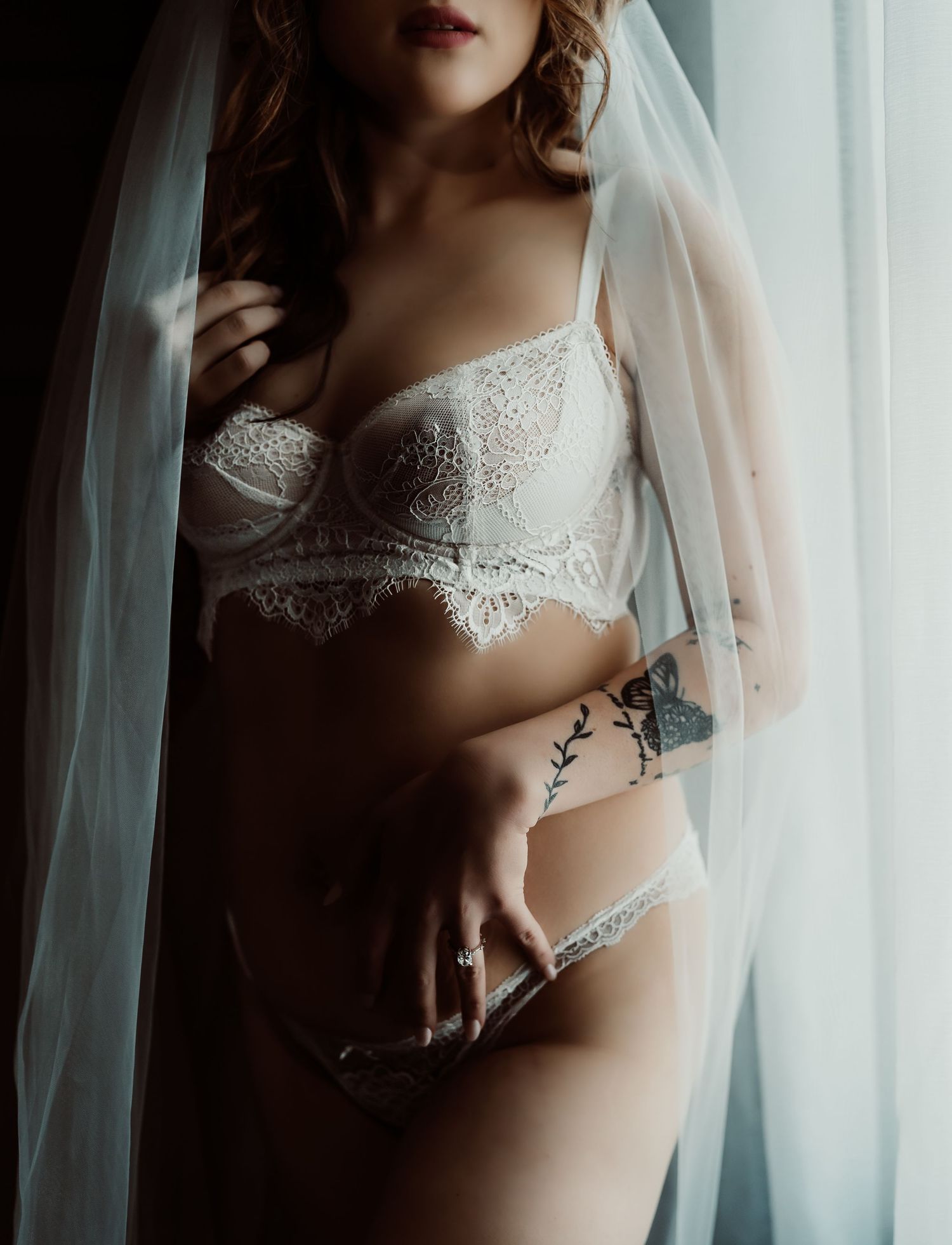Woman in white lace bridal lingerie set with veil, featuring floral tattoo on arm, posed by window.