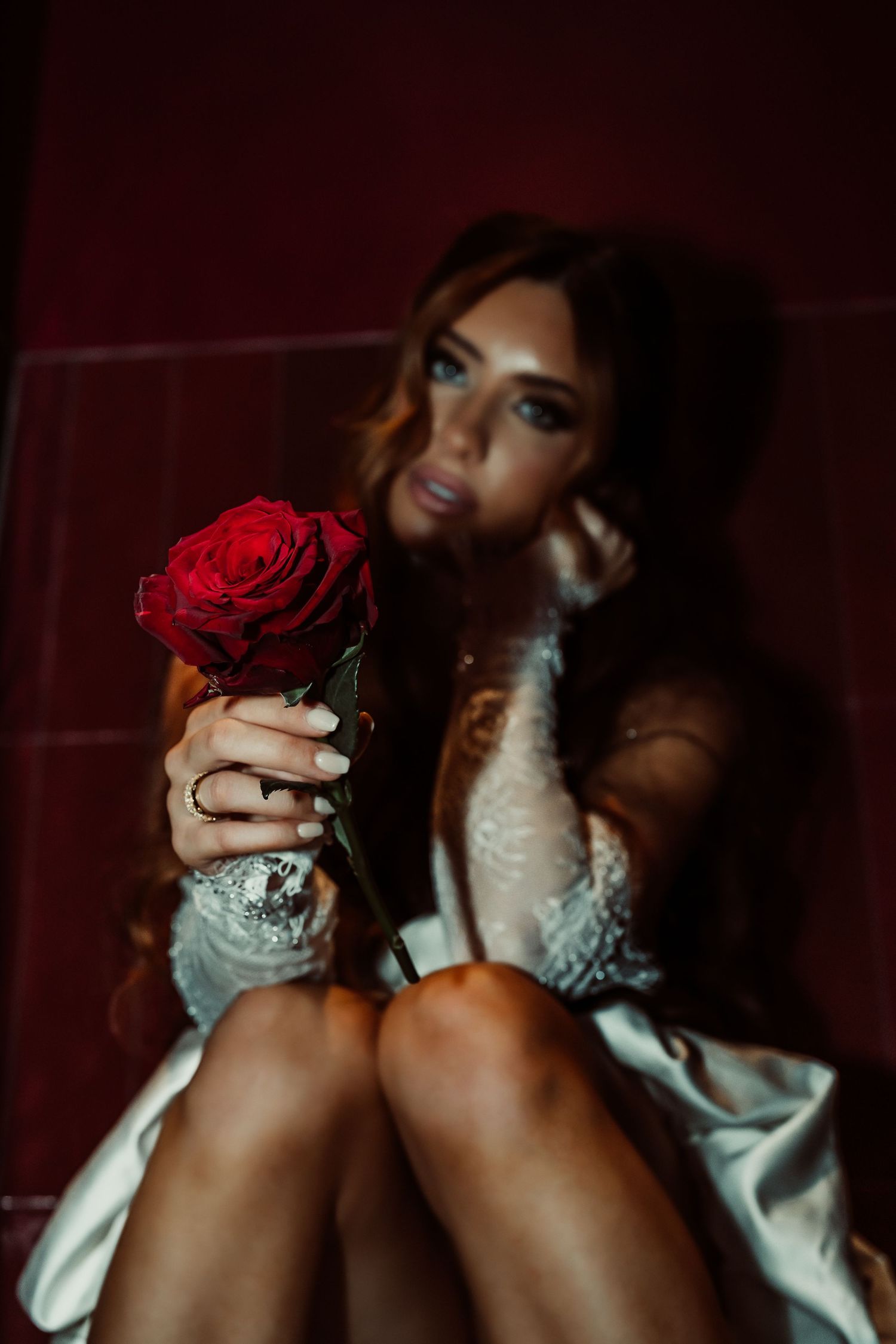 Woman in white lace robe sitting on floor holding a red rose toward camera in moody dark setting.