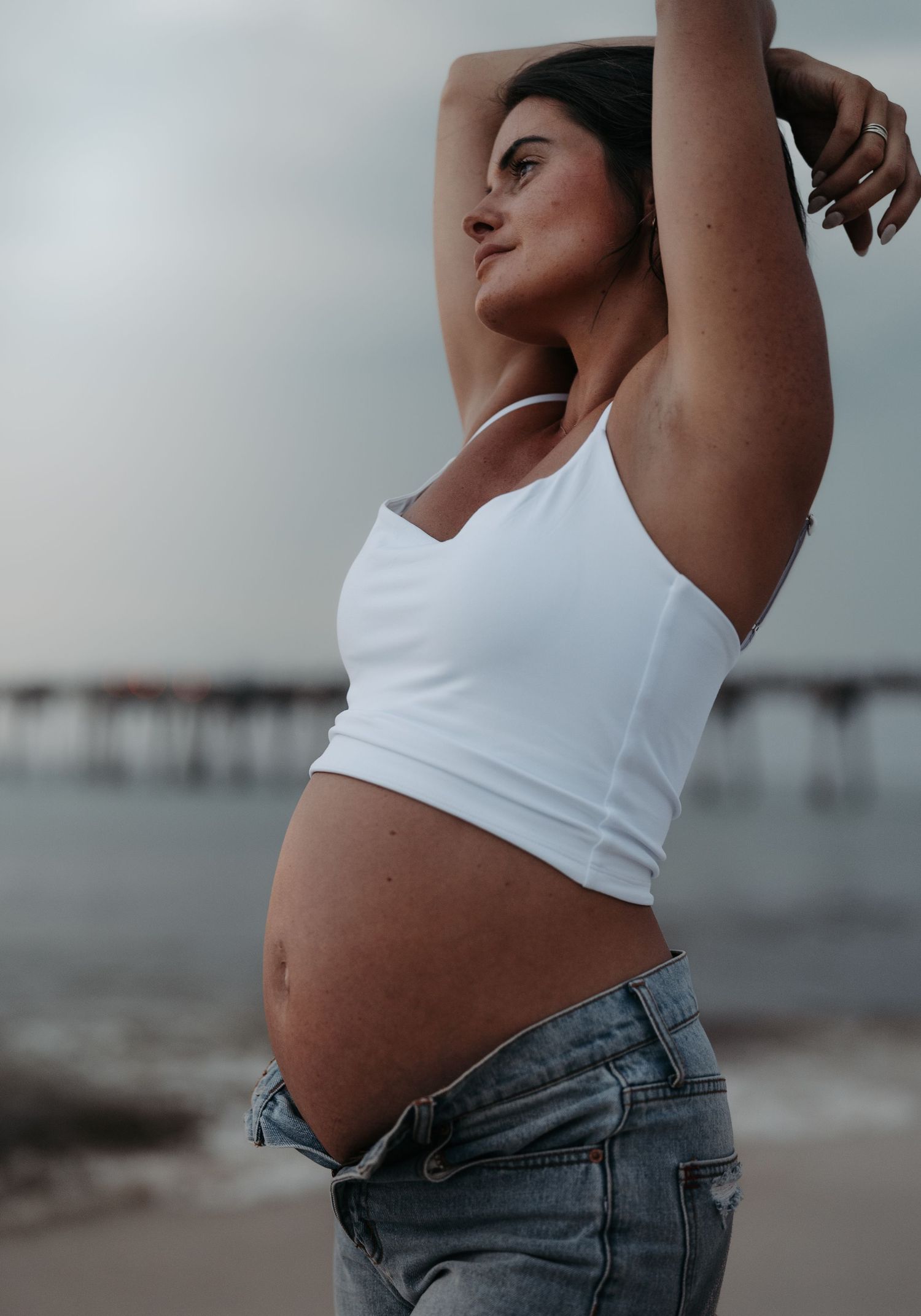 Pregnant woman in white crop top and jeans poses on beach with arms raised, showcasing baby bump near pier.