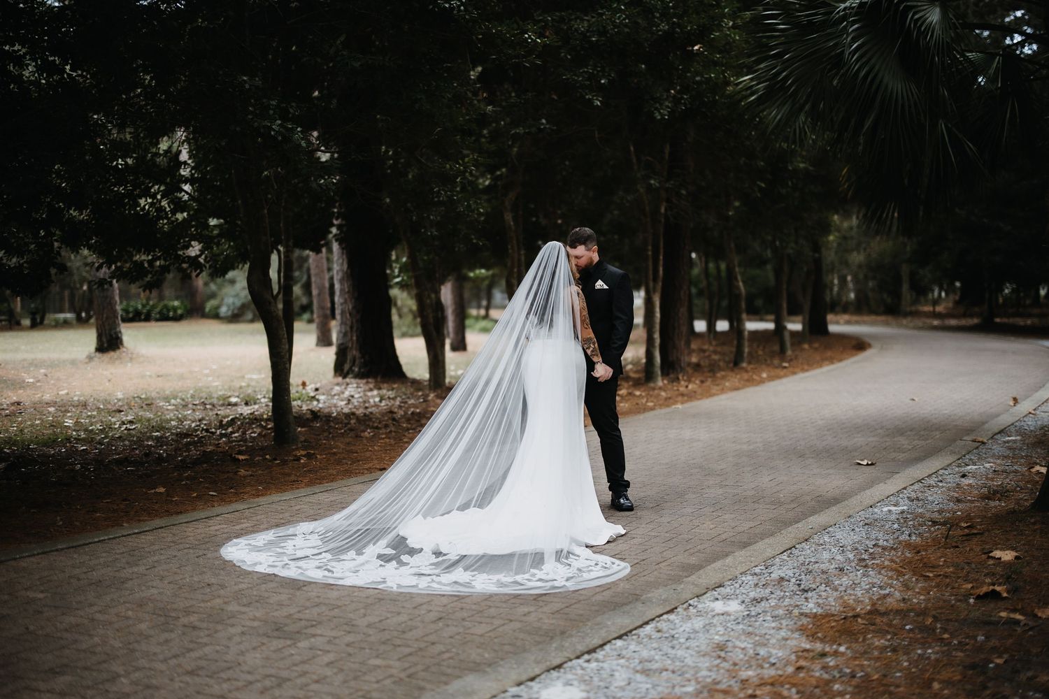 A couple in wedding attire walk down a tree-lined path with a long flowing white veil trailing behind them.