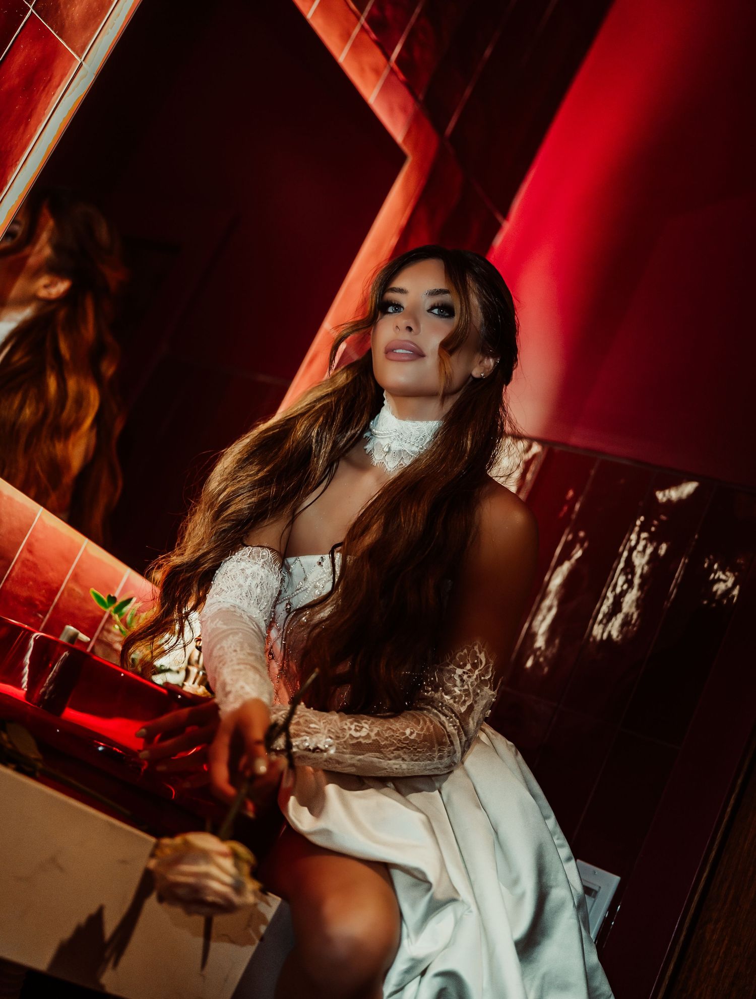 A woman in a white lace off-shoulder dress poses dramatically in a red-lit room with long flowing brunette hair.
