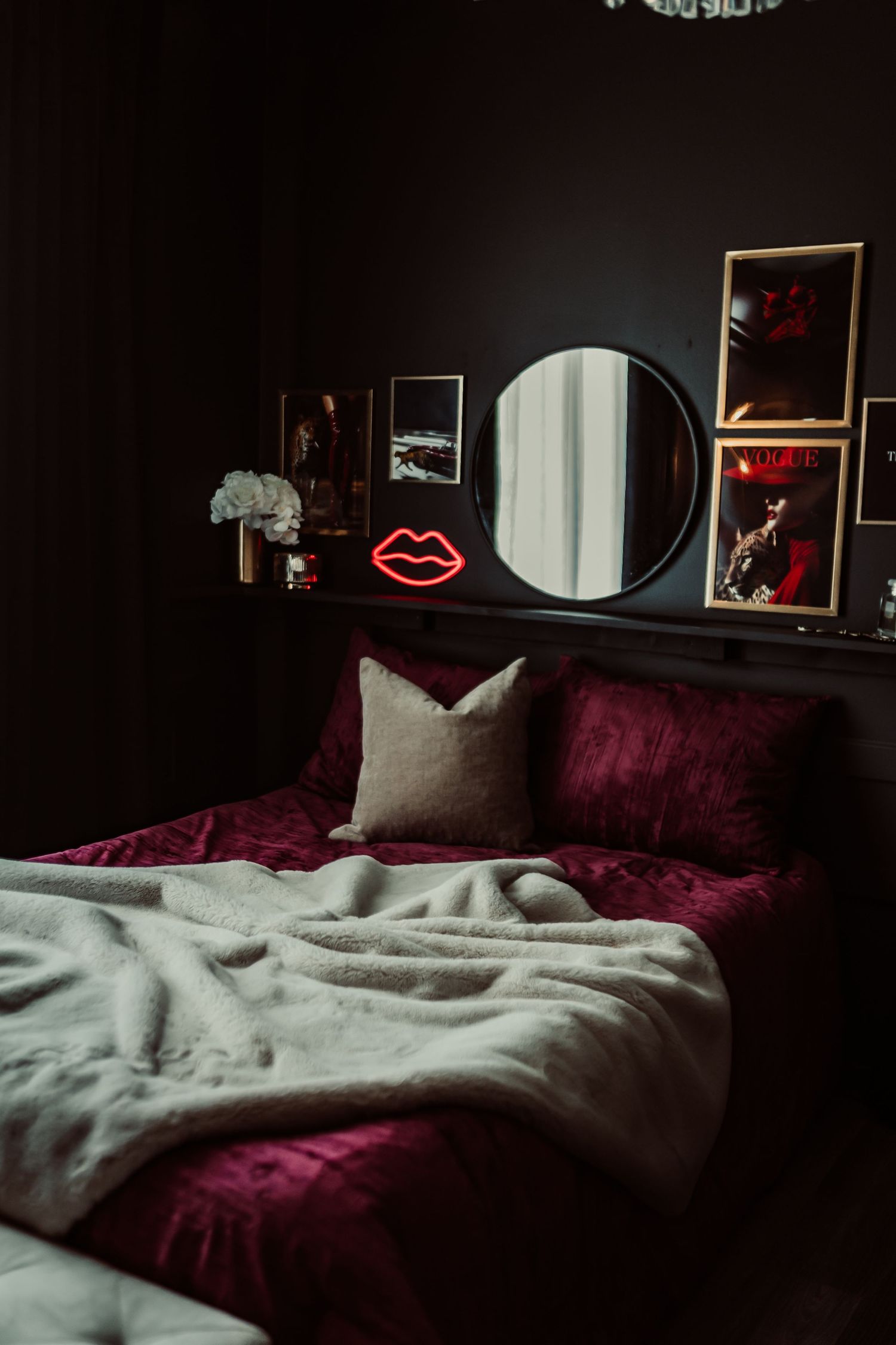 Dark moody bedroom with crimson velvet bedding, round mirror, neon lips sign, and framed wall art above the headboard.