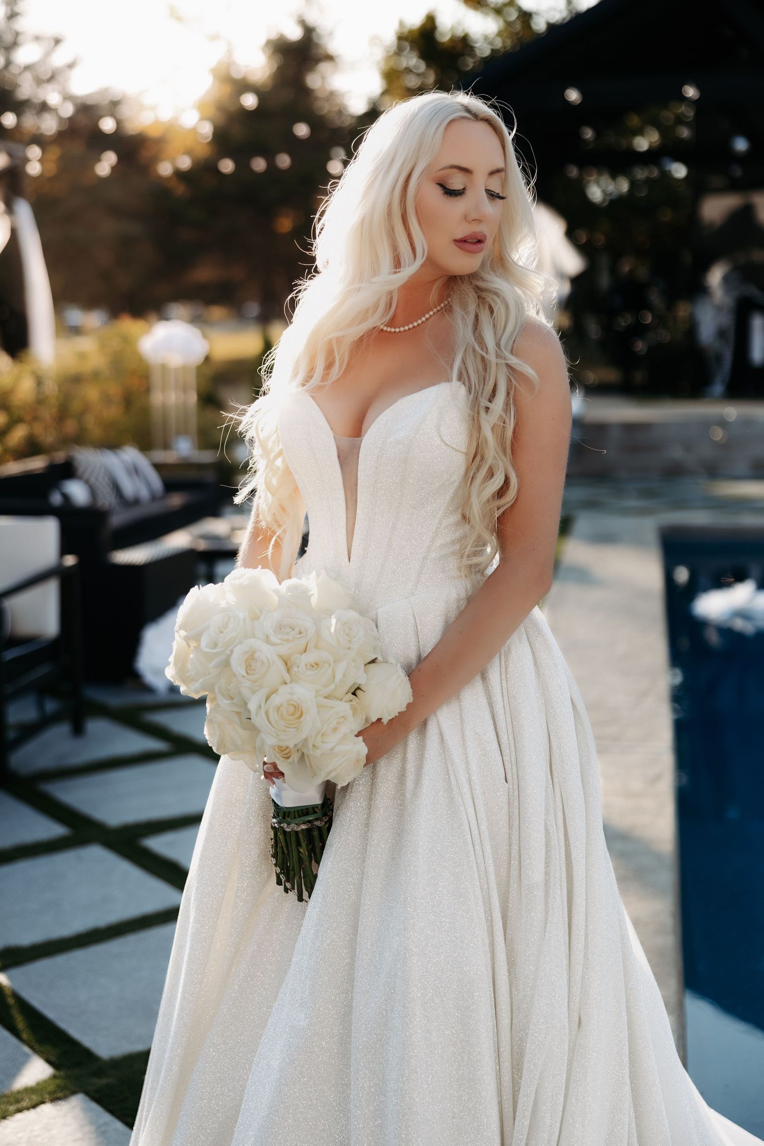 Elegant bride in flowing white gown holds bouquet of white roses during golden hour sunset photoshoot.