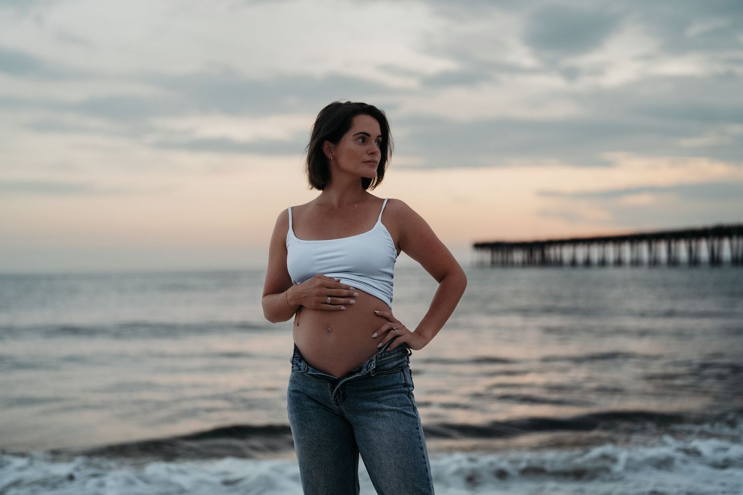 Pregnant woman in white crop top and jeans poses on beach at sunset with pier in background.