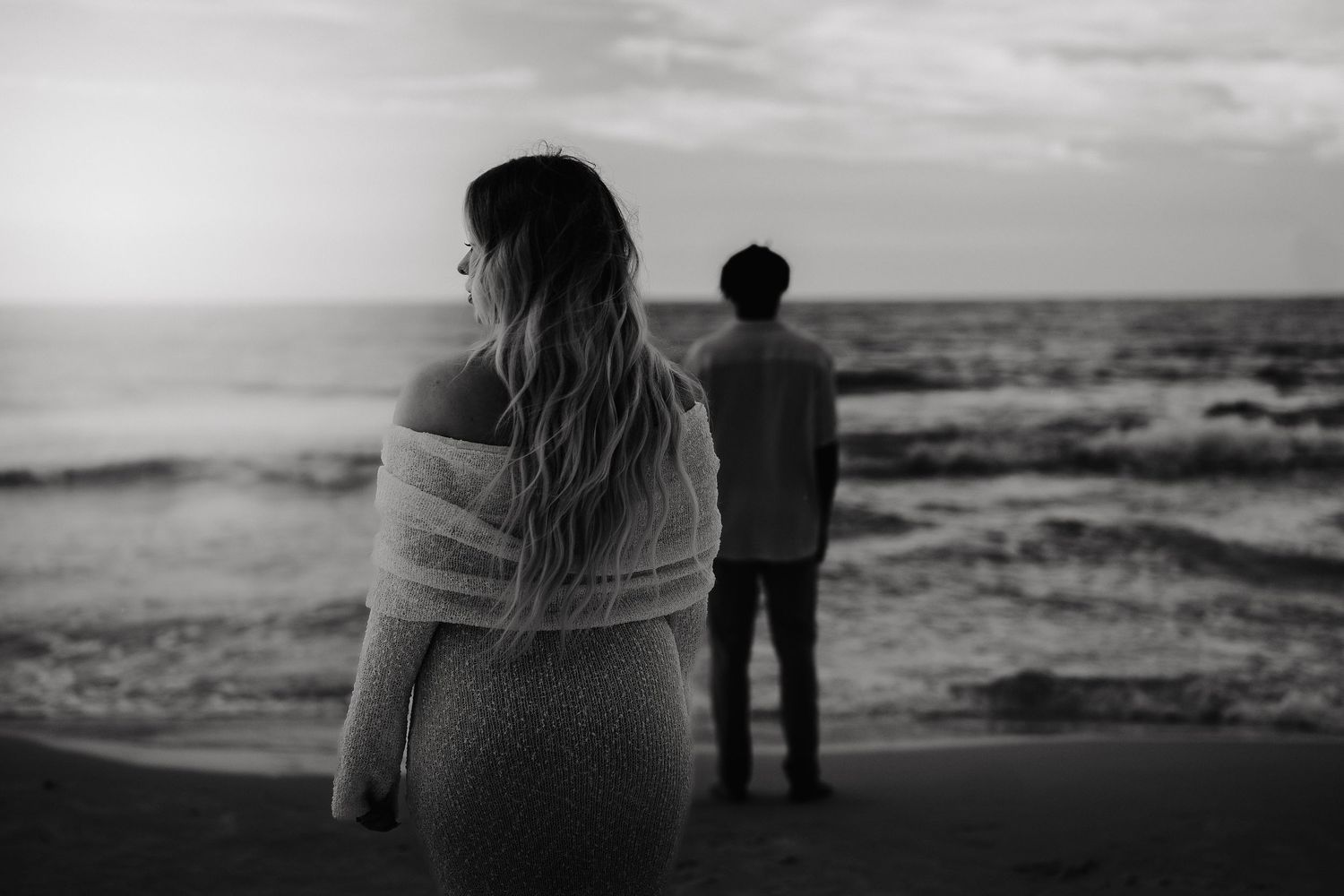 Black and white photo of a couple standing apart on a beach at sunset, woman in foreground, man near ocean waves.