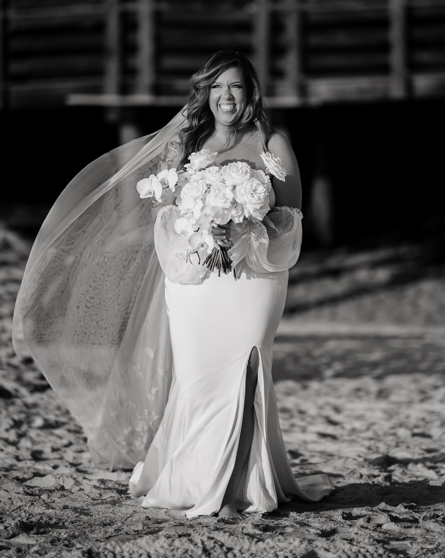 A bride in a flowing white wedding dress sits gracefully on the beach during a seaside photoshoot.