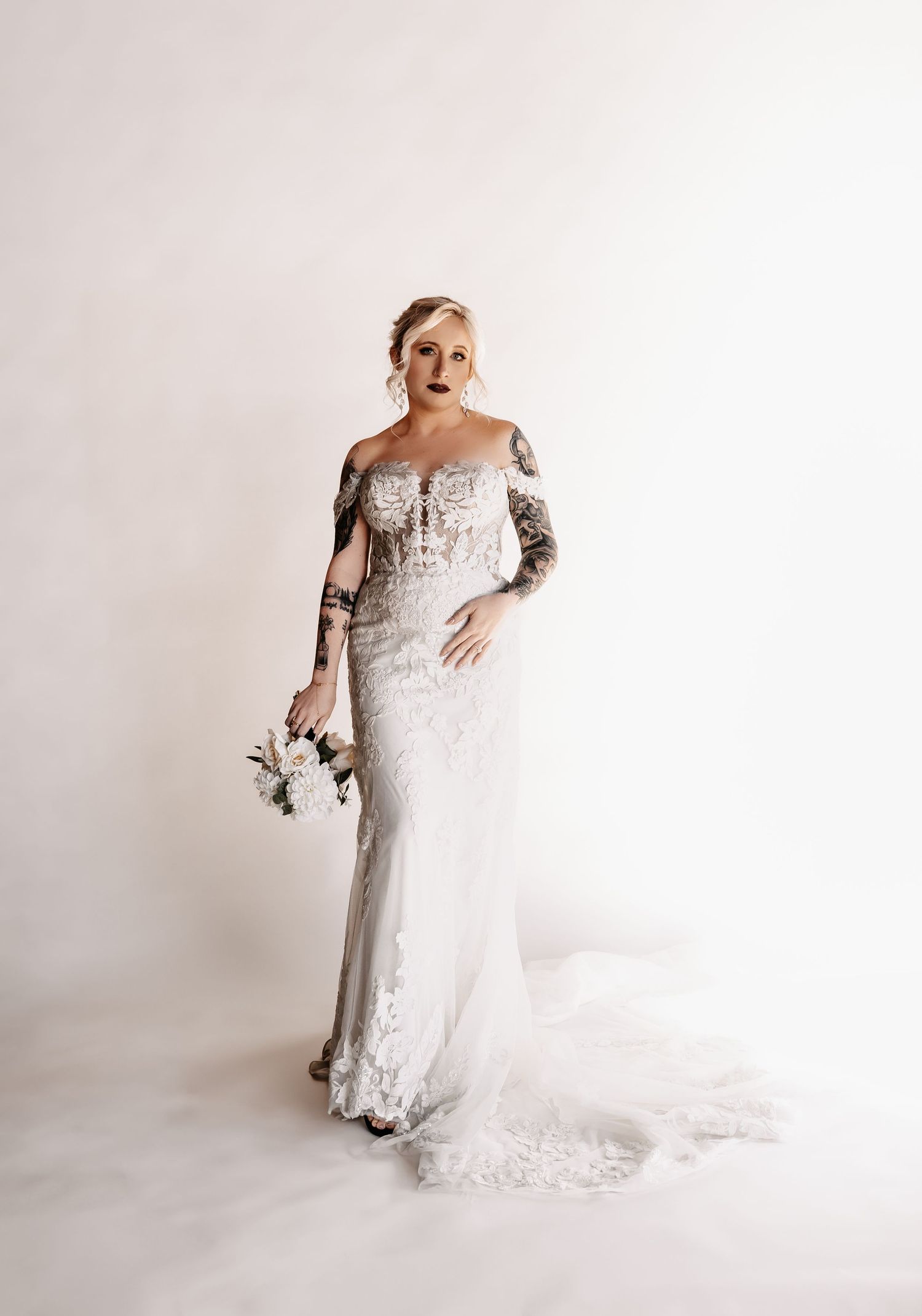Tattooed bride in off-shoulder lace wedding gown with train holds white bouquet against white background.
