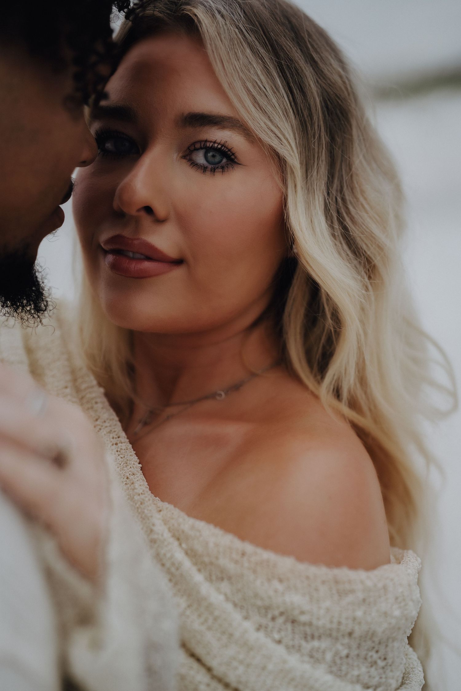 Close-up portrait of a blonde woman with blue eyes wearing an off-shoulder cream sweater and delicate necklace.