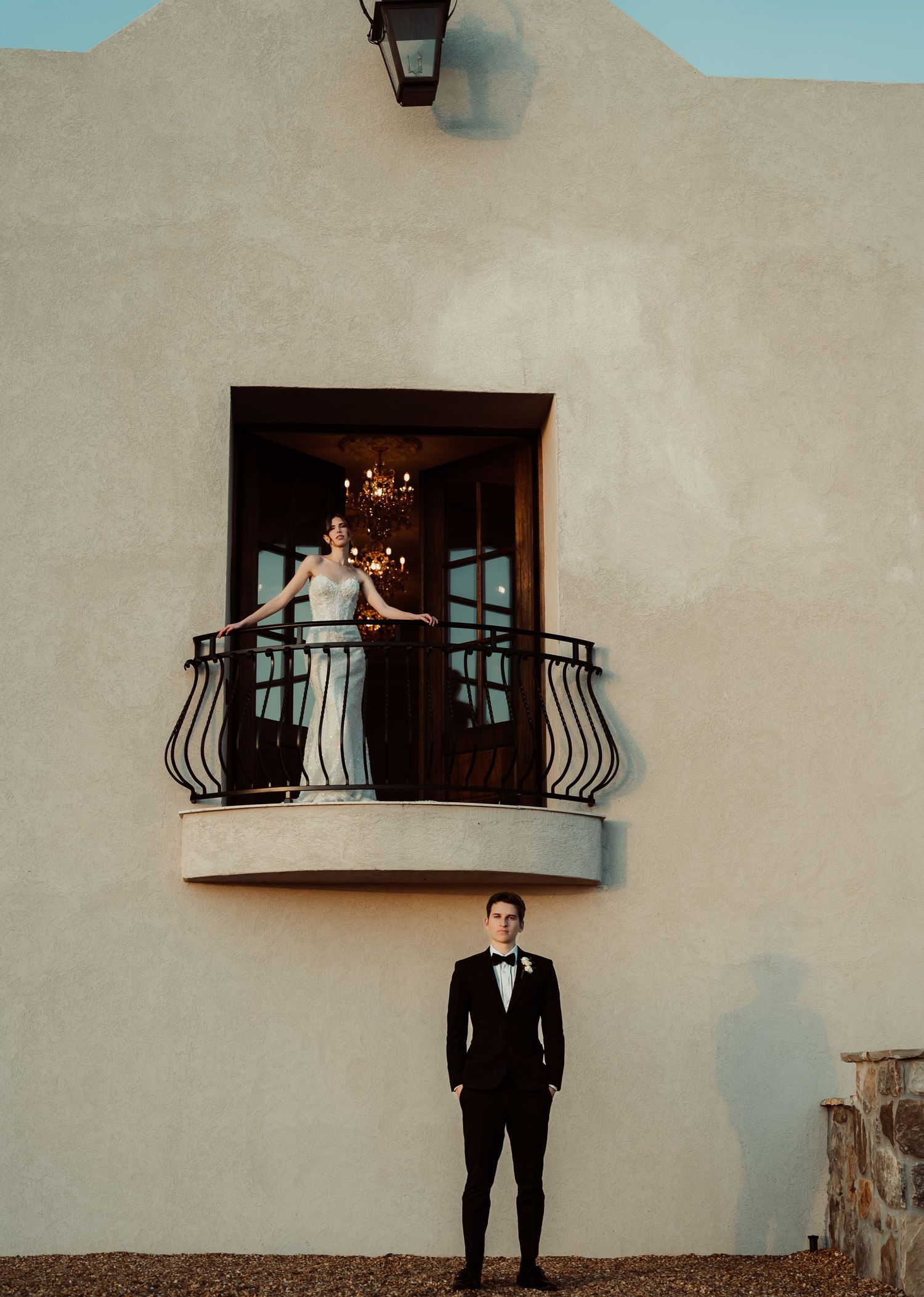 Bride on balcony with groom standing below in tuxedo at elegant wedding venue with stucco walls.