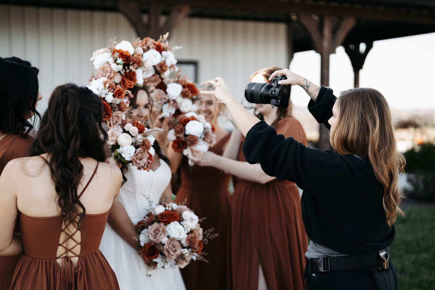 A group of bridesmaids in burgundy dresses stand together holding white and rust-colored floral bouquets at a wedding.