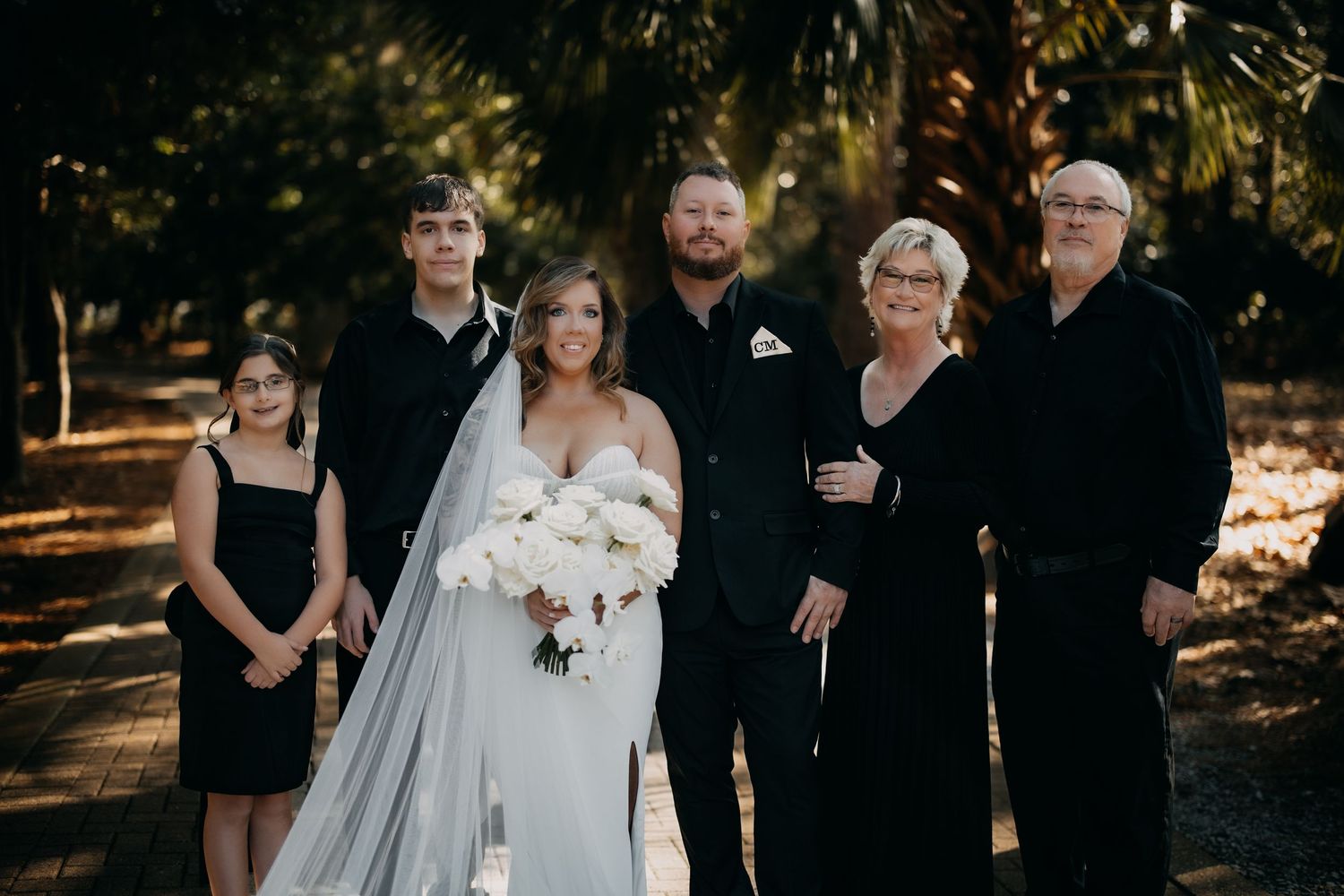 A wedding party stands together in formal black and white attire under trees.