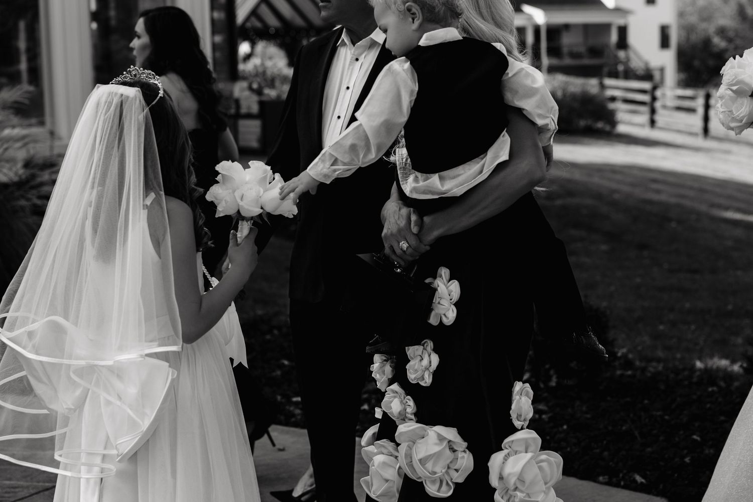 Black and white wedding photo of bride in veil holding flowers with guests in floral dress outdoors.