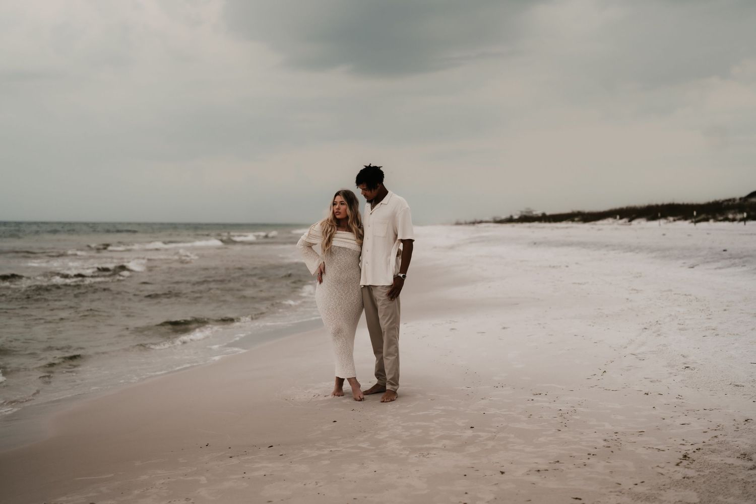 A couple in white outfits walks romantically on a sandy beach with gentle waves under a cloudy sky.