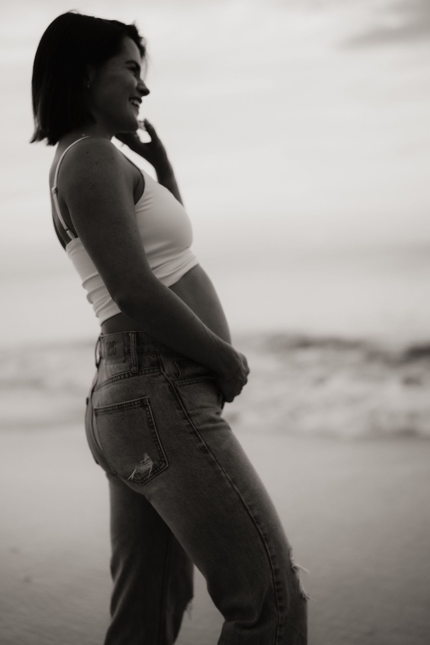 Black and white photo of a smiling pregnant woman in a crop top and jeans standing on a beach.