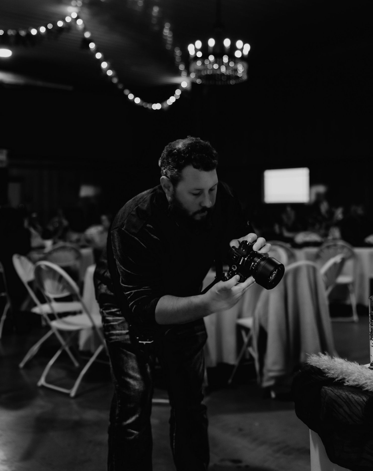 A photographer reviews photos on their camera at an indoor event with string lights and folding chairs in the background.