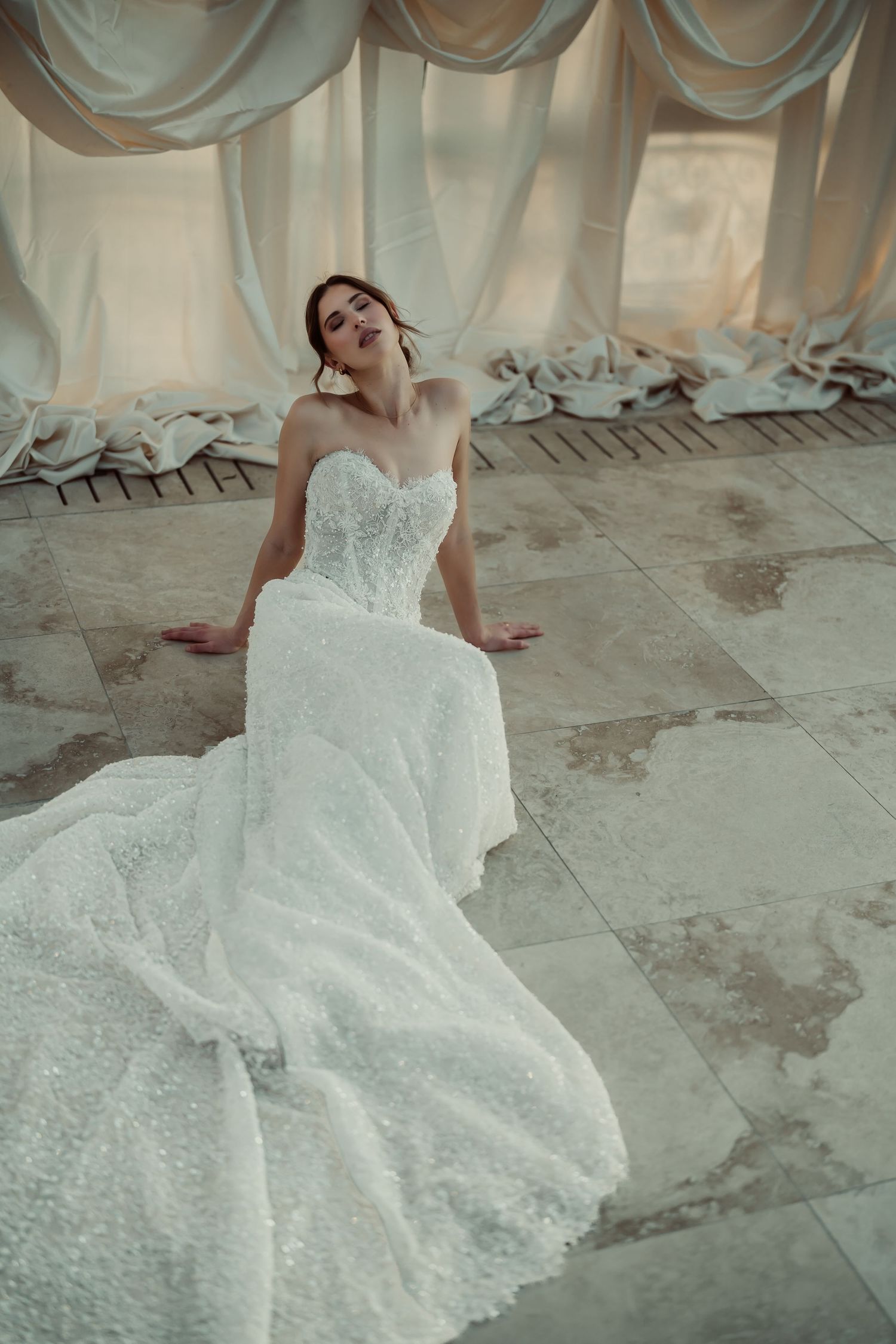 Bride in strapless beaded white ball gown with long train sits gracefully on marble floor amid draped white curtains.