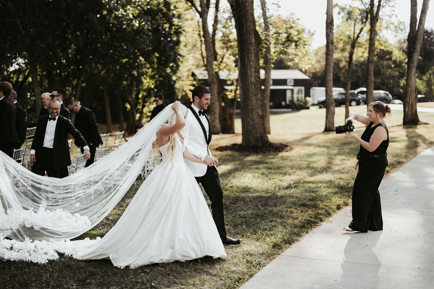 A wedding party walks along a tree-lined path with a flowing white veil trailing behind the bride's gown.