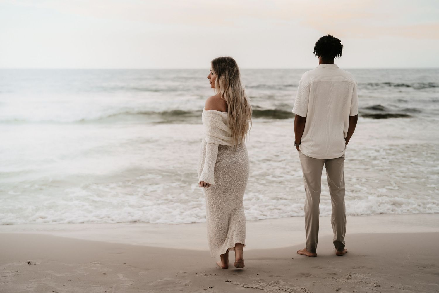 Couple standing together on a sandy beach facing ocean waves, woman in white dress and man in casual white outfit.