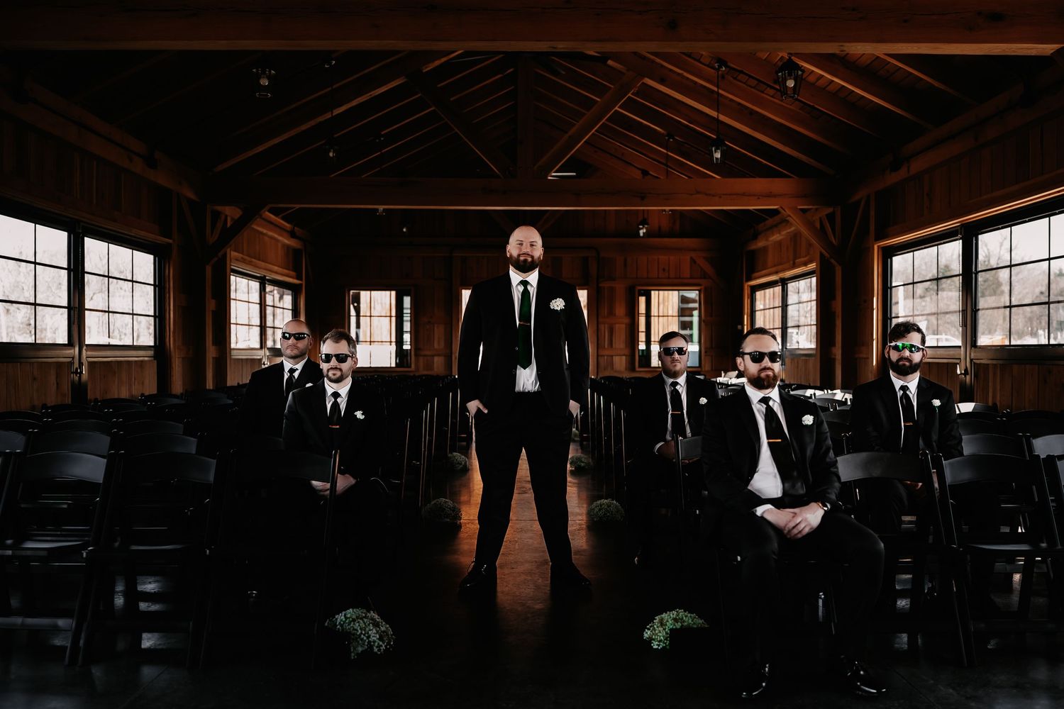 Groom stands confidently among groomsmen in a rustic wooden barn venue, all dressed in elegant black suits.