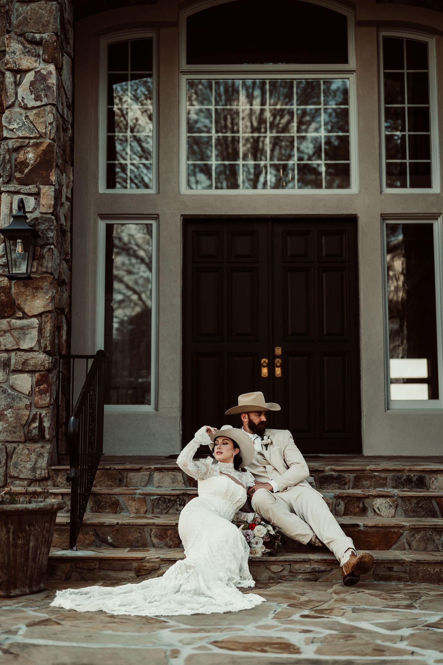 Bride and groom pose on stone steps in white gown and tan cowboy hat suit outside rustic venue.