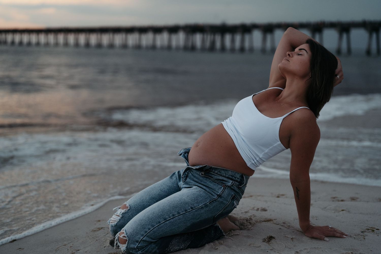 Woman in white crop top and jeans posing on sandy beach at sunset with pier in background.