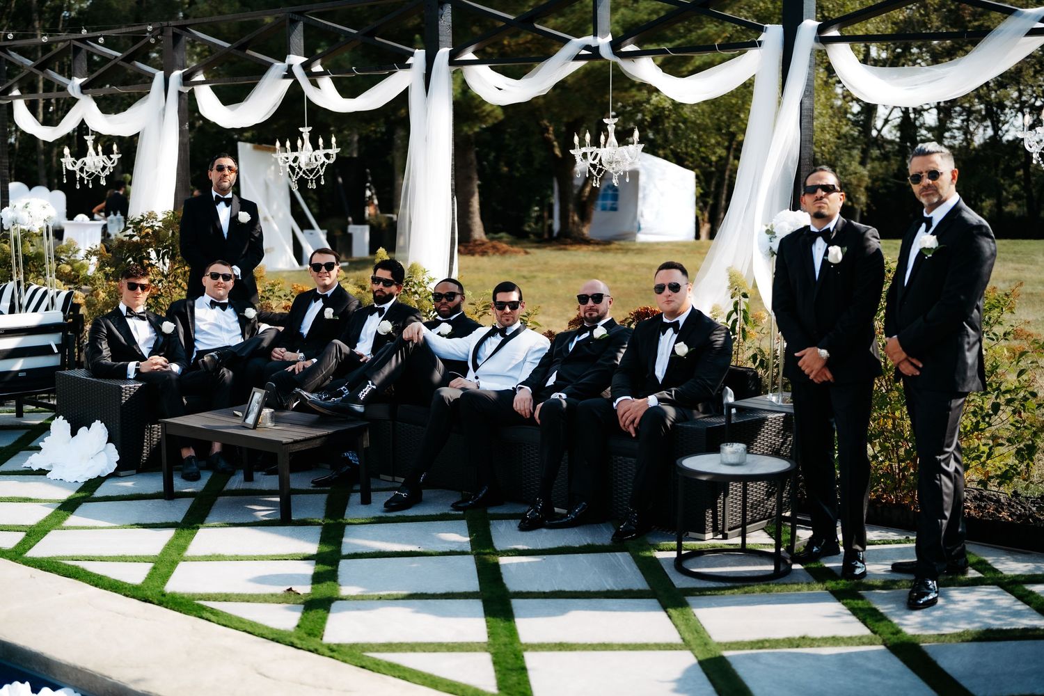 Wedding party members in black tuxedos sit together on outdoor seating during an elegant ceremony with white draping decor.