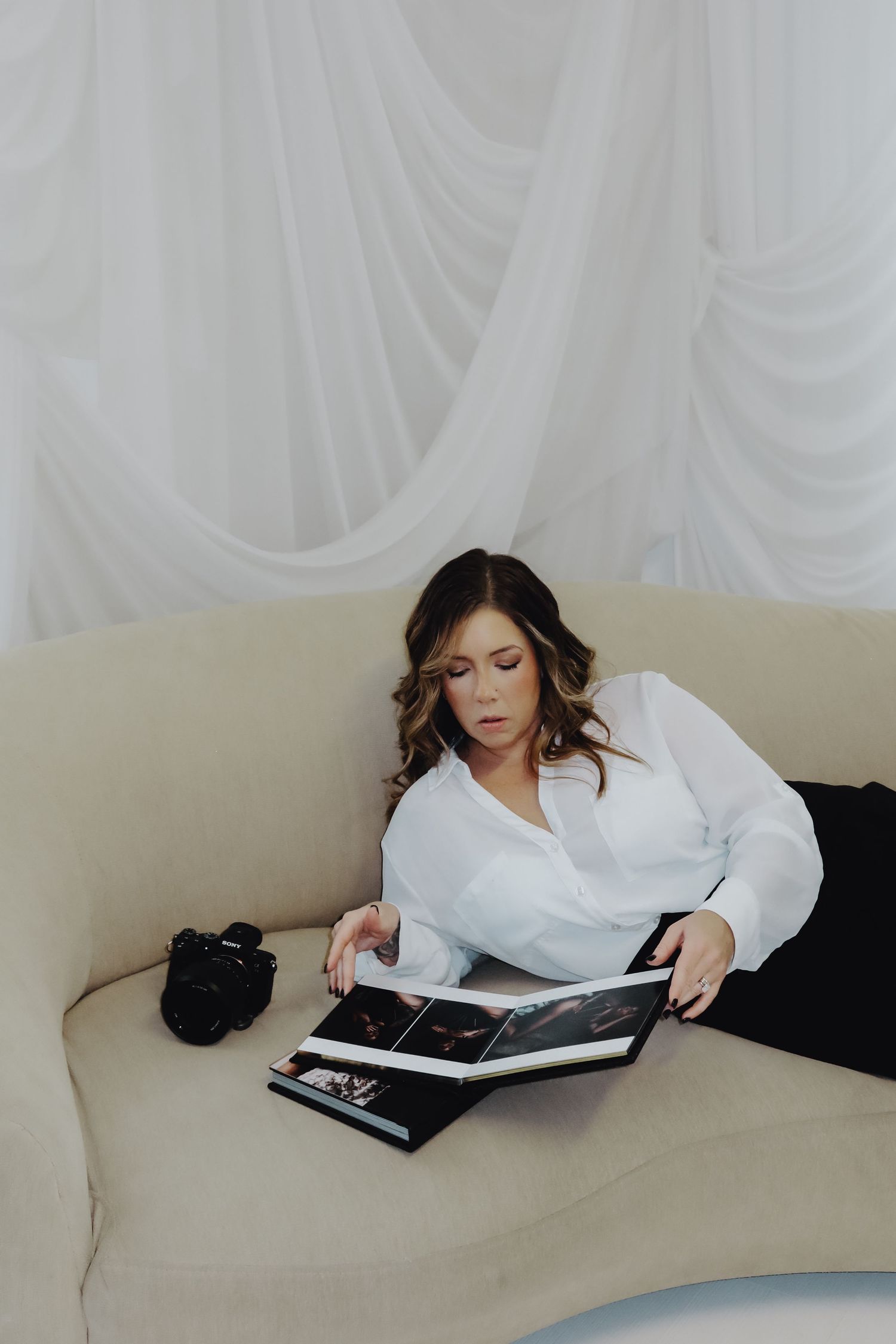 Woman in white blouse reviews photo album on cream sofa with camera nearby, set against elegant white draped backdrop.