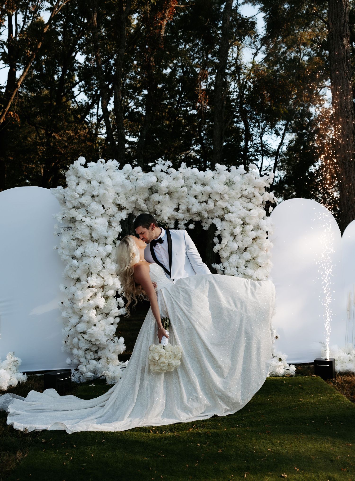 A bride in a flowing white gown poses gracefully on grass against a backdrop of white balloon arches in an outdoor setting.