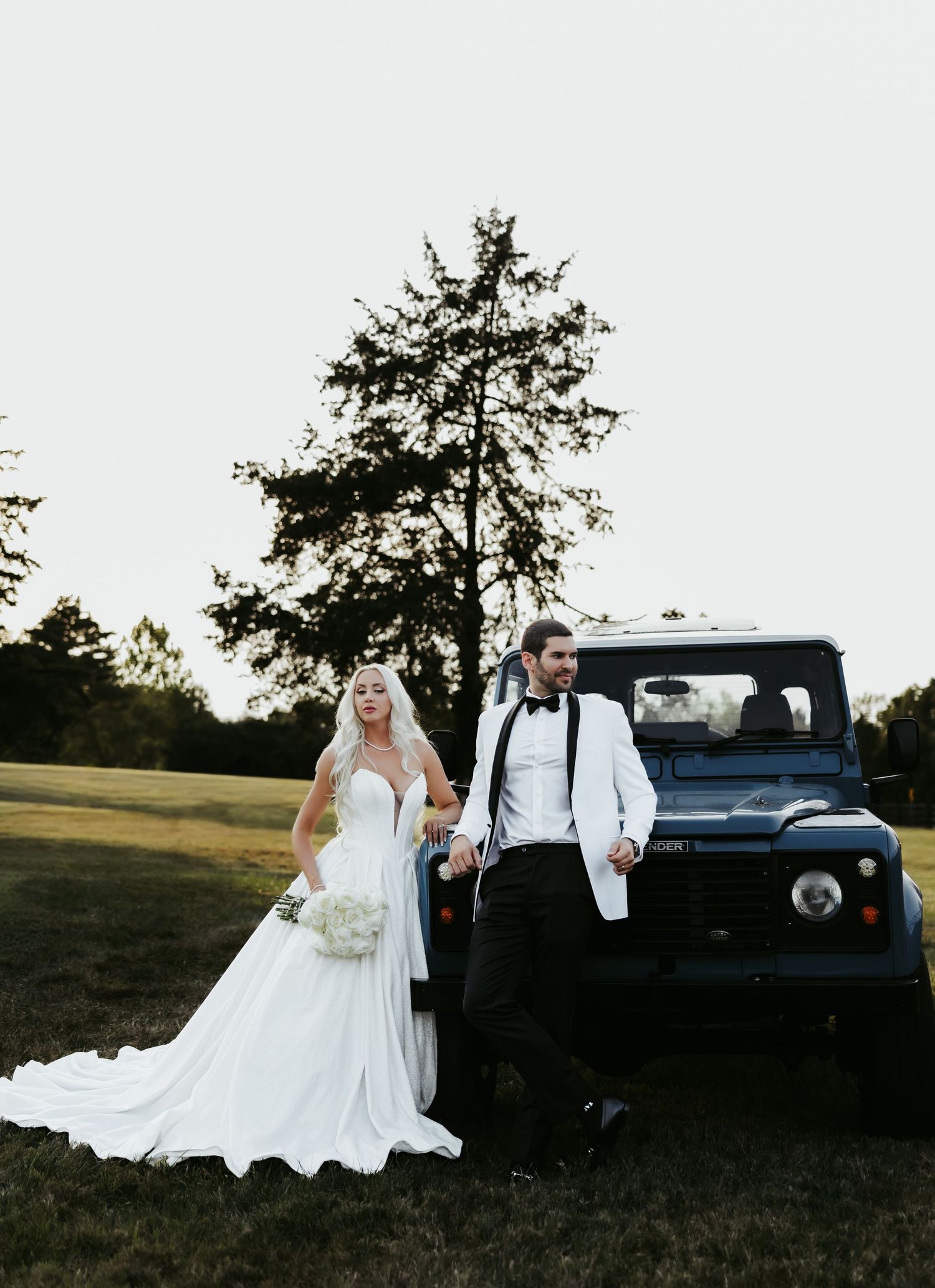 Newlyweds pose beside vintage blue truck in rustic outdoor setting with tall evergreen trees.