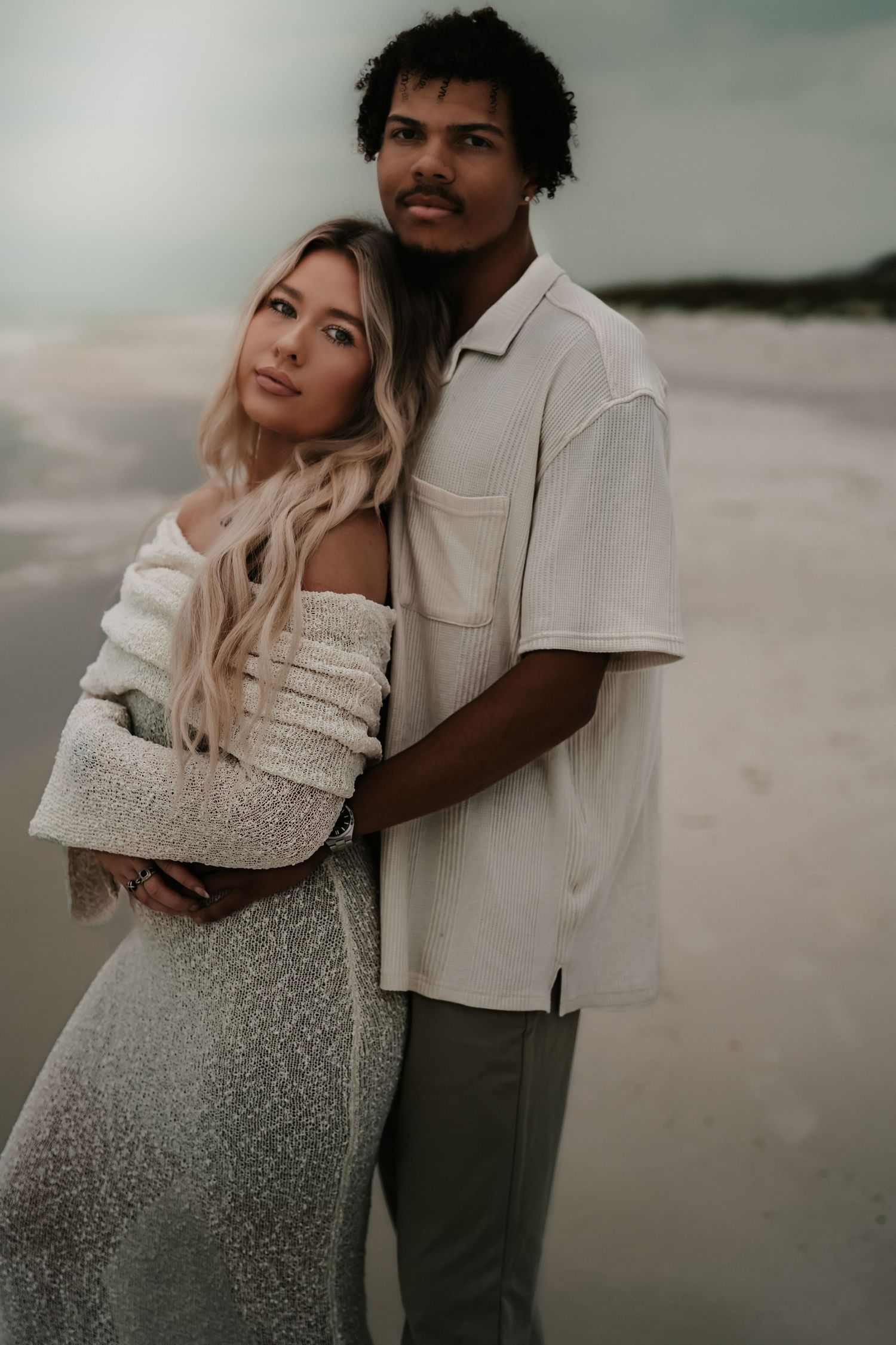 A couple embraces on a beach, woman in white off-shoulder dress, man in cream shirt, posing for romantic portrait.