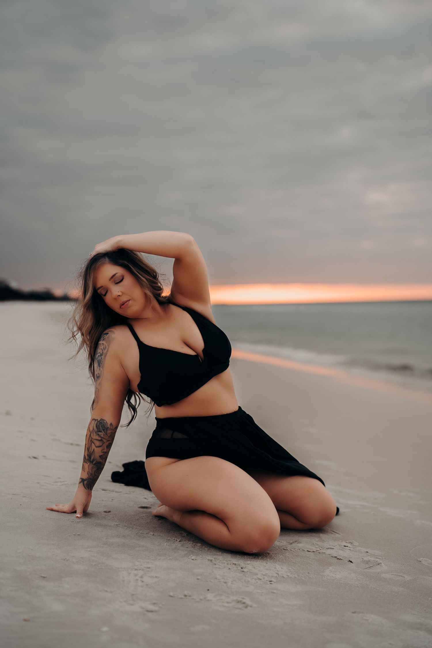 Woman in black bikini top and skirt kneeling on sandy beach at sunset with dramatic cloudy sky.