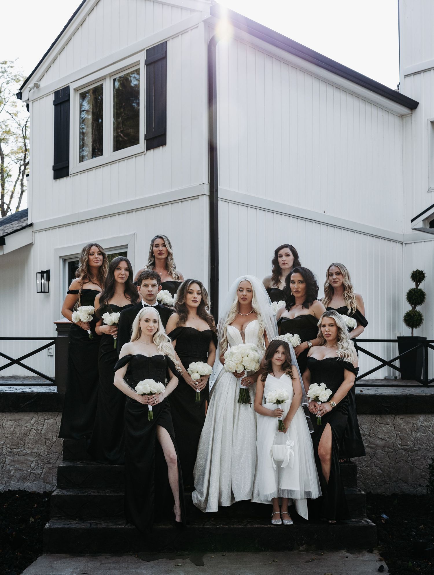 Wedding party in black and white dresses pose together against white building exterior with stone foundation.
Stone Haven