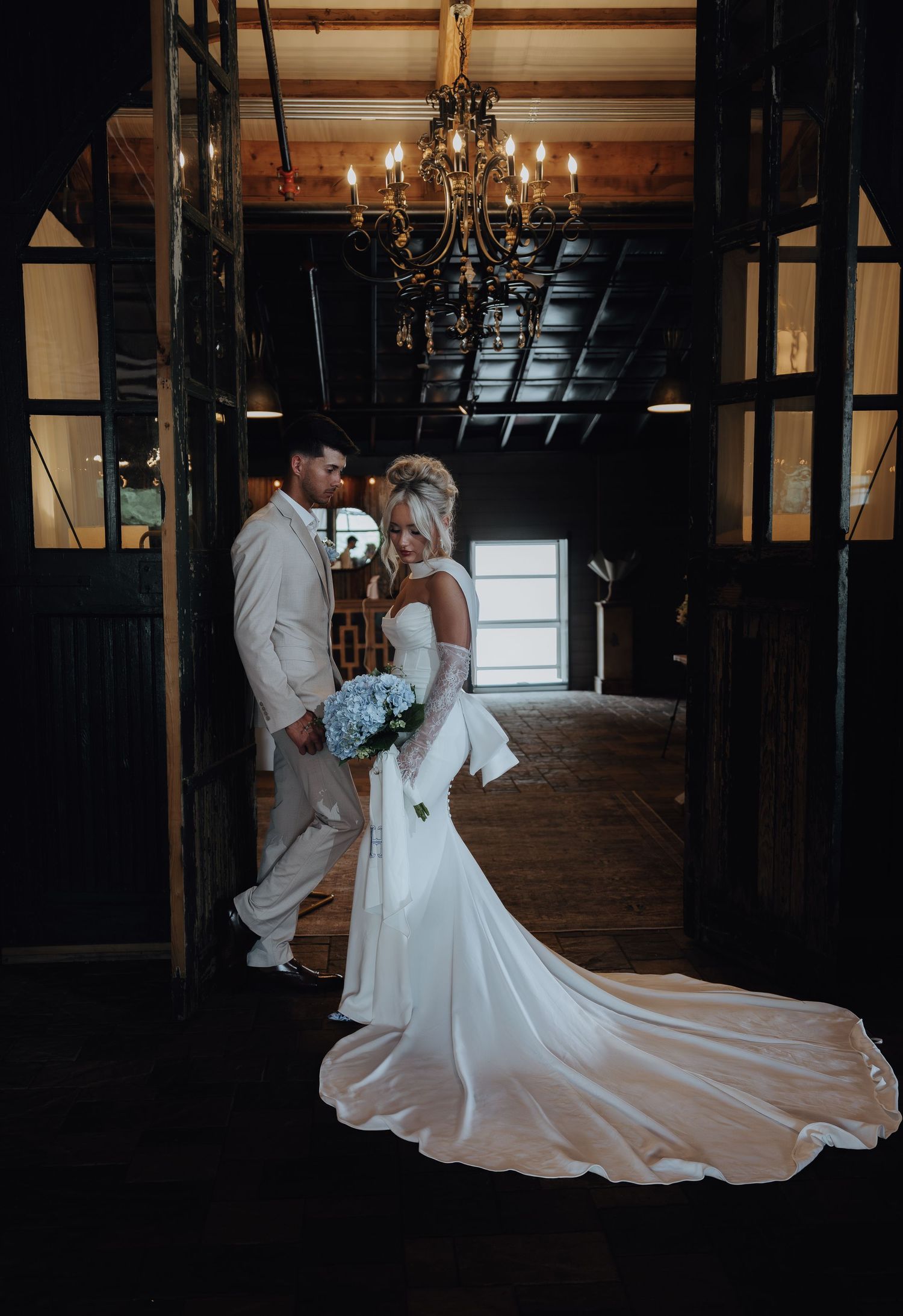 Bride in white gown with train holds blue bouquet beside groom in tan suit under chandelier in rustic barn.