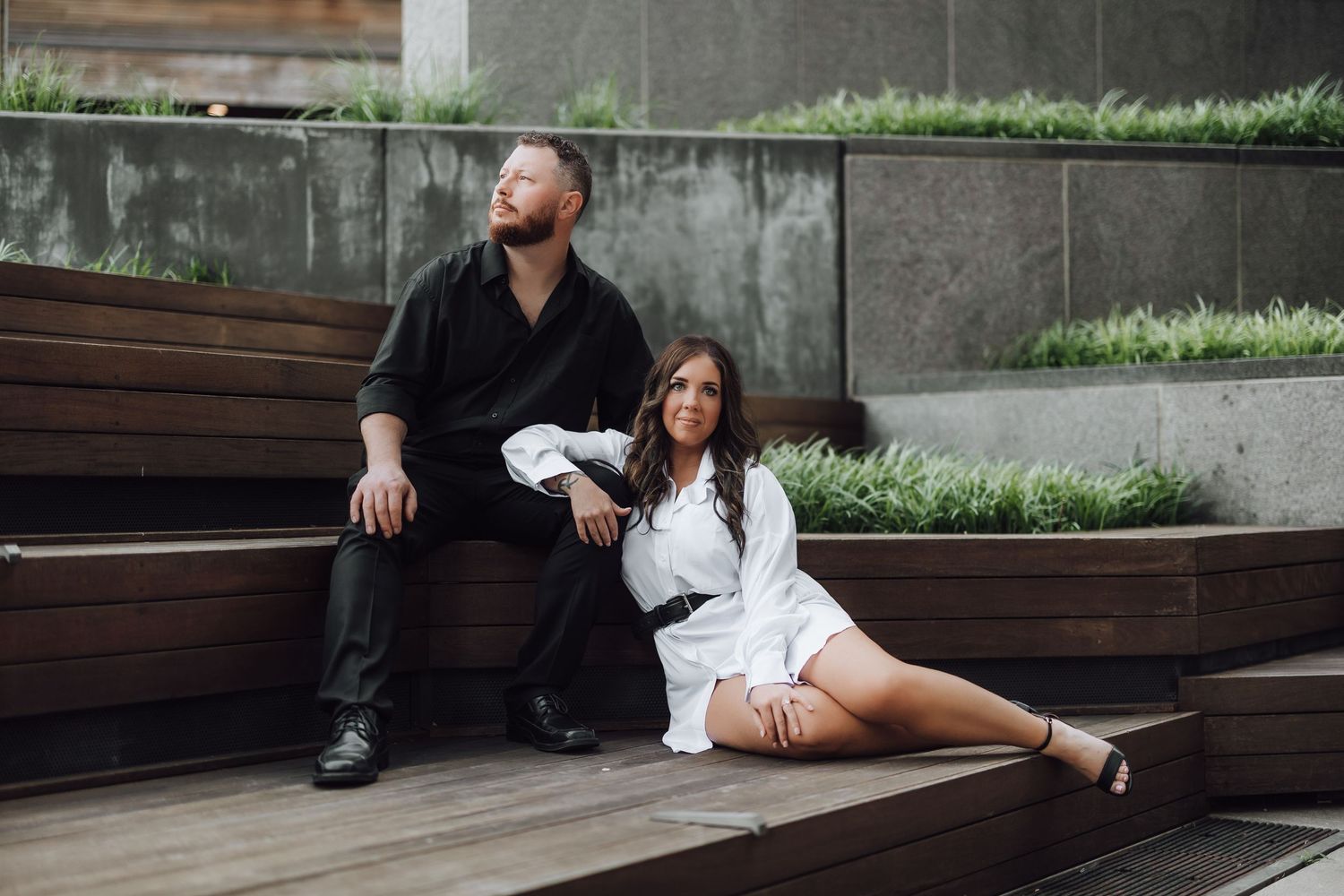 Couple sits together on wooden bench in modern outdoor courtyard with concrete walls and greenery.