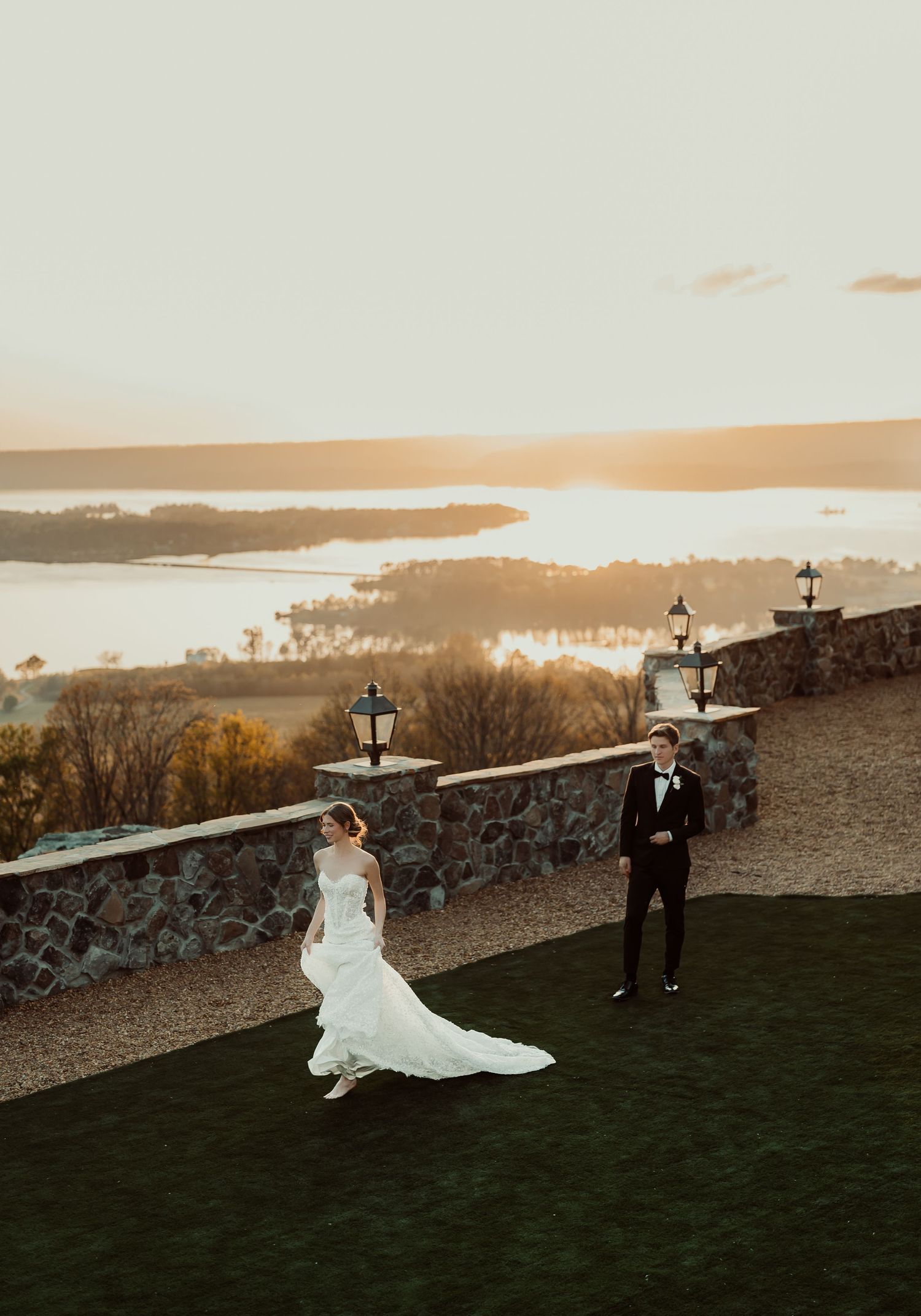 Bride and groom stand on a stone terrace at sunset with a scenic lake and golden sky backdrop.