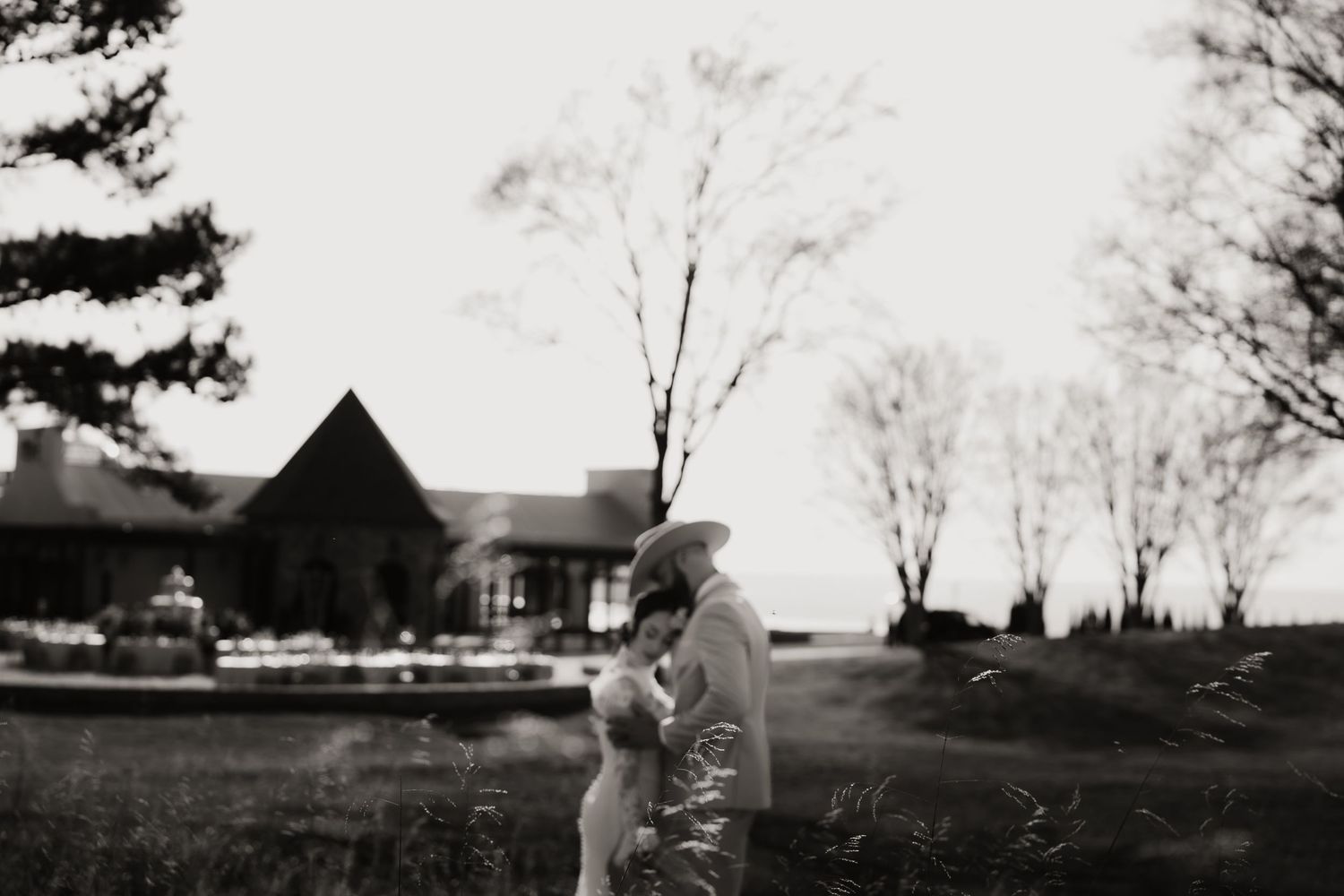 Black and white photo of a couple embracing outdoors near a rustic building surrounded by bare trees.