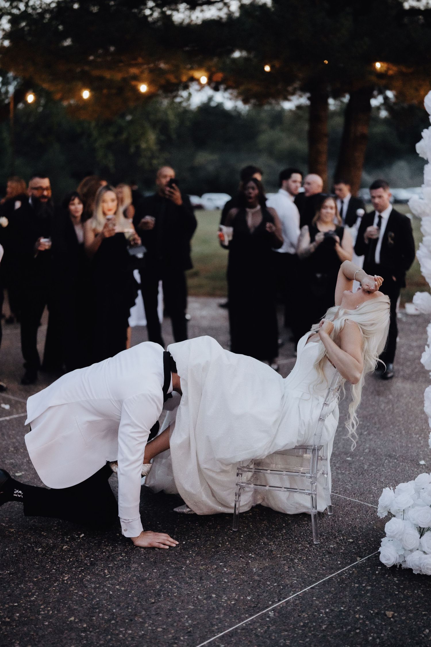 Groom retrieves garter from bride seated on chair at outdoor wedding reception with guests in black attire.
