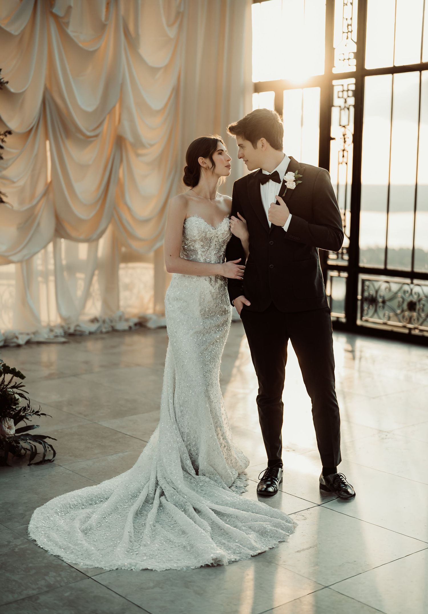 Elegant bride in beaded mermaid gown and groom in black tuxedo share a romantic moment in a sunlit ballroom.