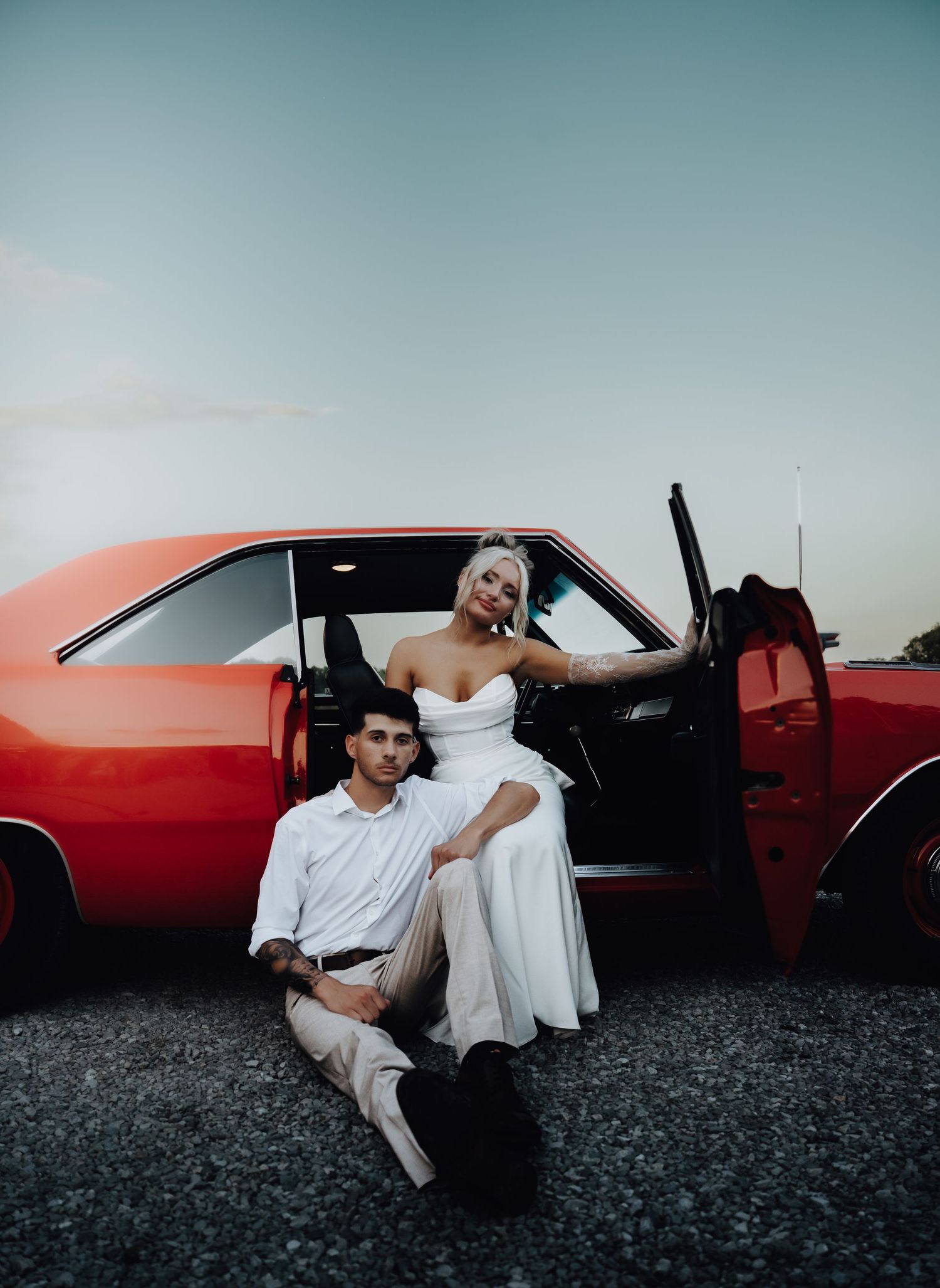 A couple poses by a classic red muscle car at dusk, woman in white dress sitting on door, man seated on ground.