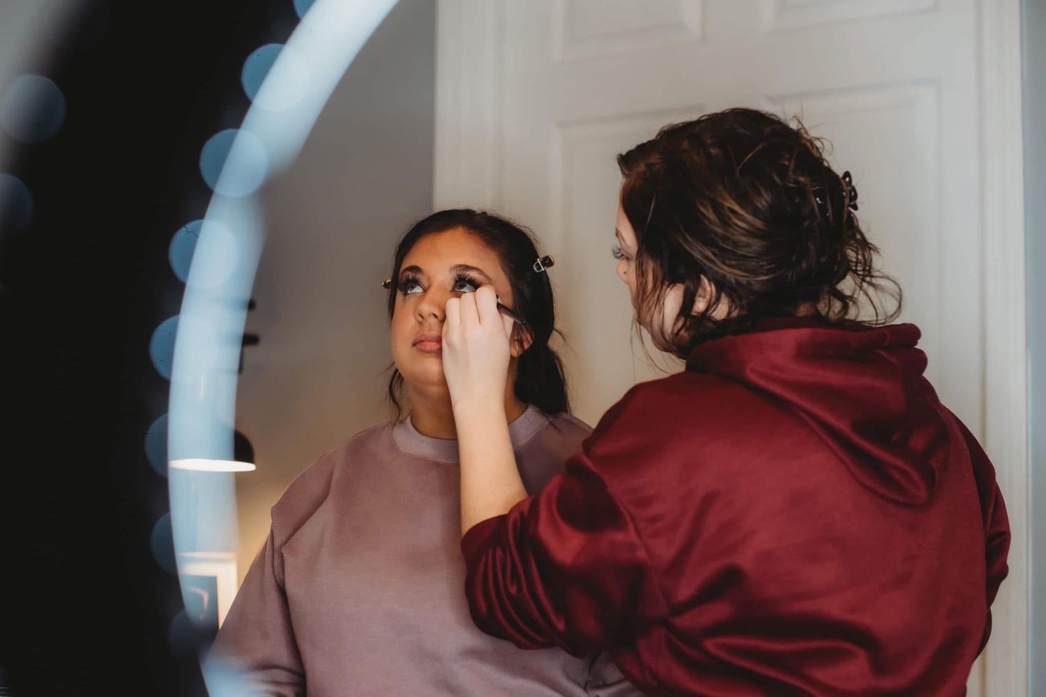Makeup artist applying eye makeup to a woman in a pink sweatshirt using a ring light.