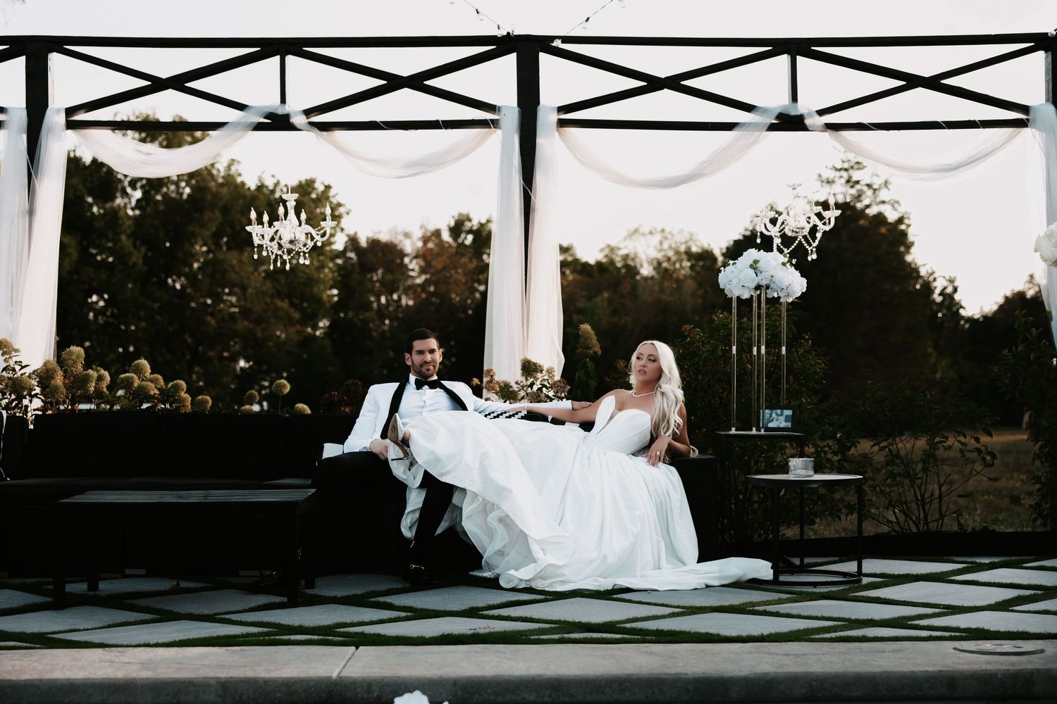 A couple relaxes on a black and white checkered dance floor under an elegant outdoor pavilion at dusk.