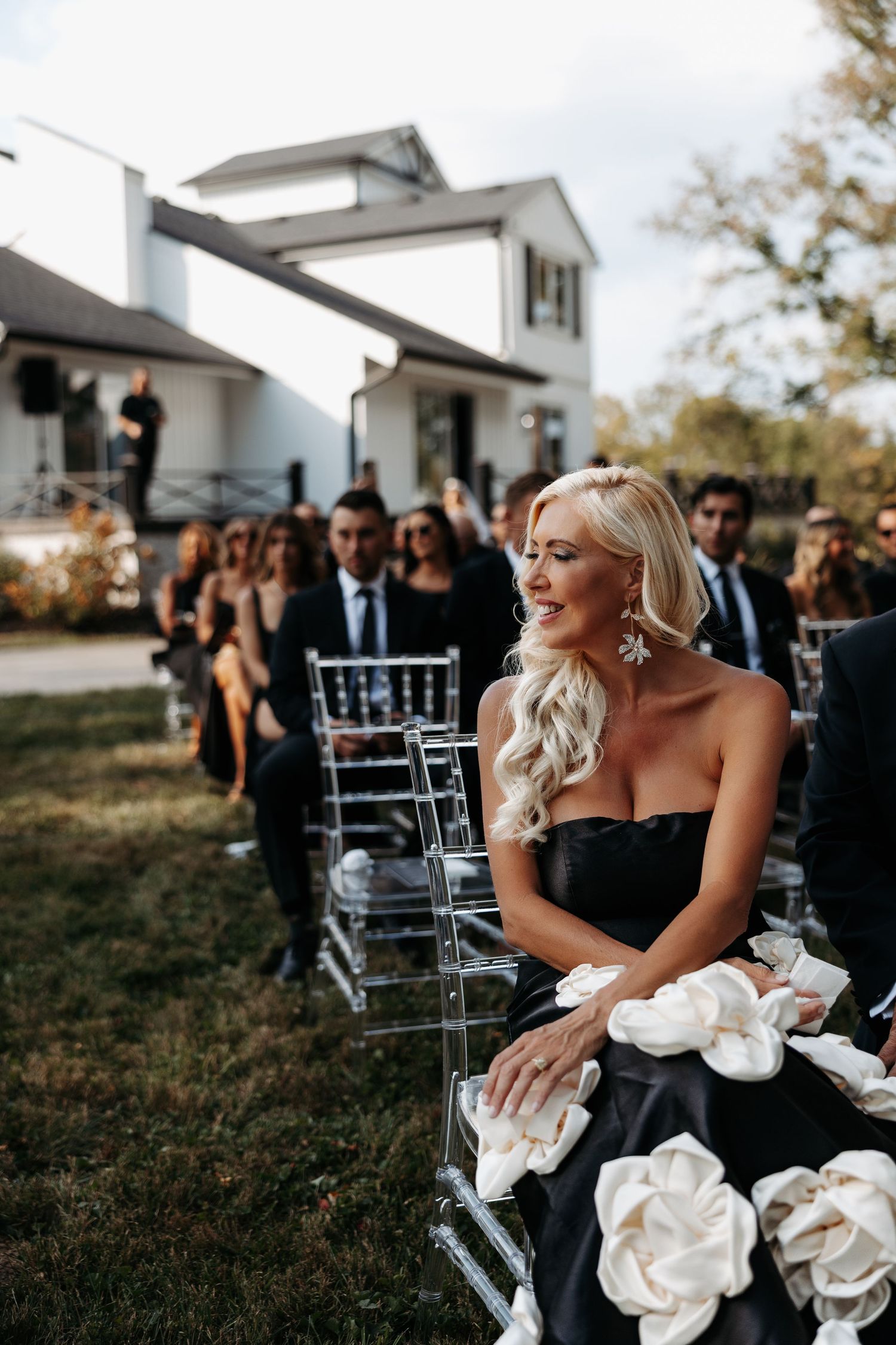 Wedding party laughs together during outdoor celebration with clear chairs and white floral arrangements.