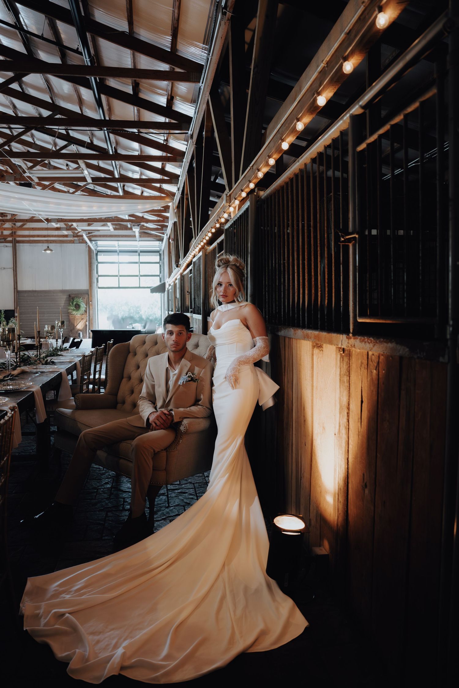 Bride in white gown with train stands beside groom in tan suit at elegant barn wedding venue with string lights.