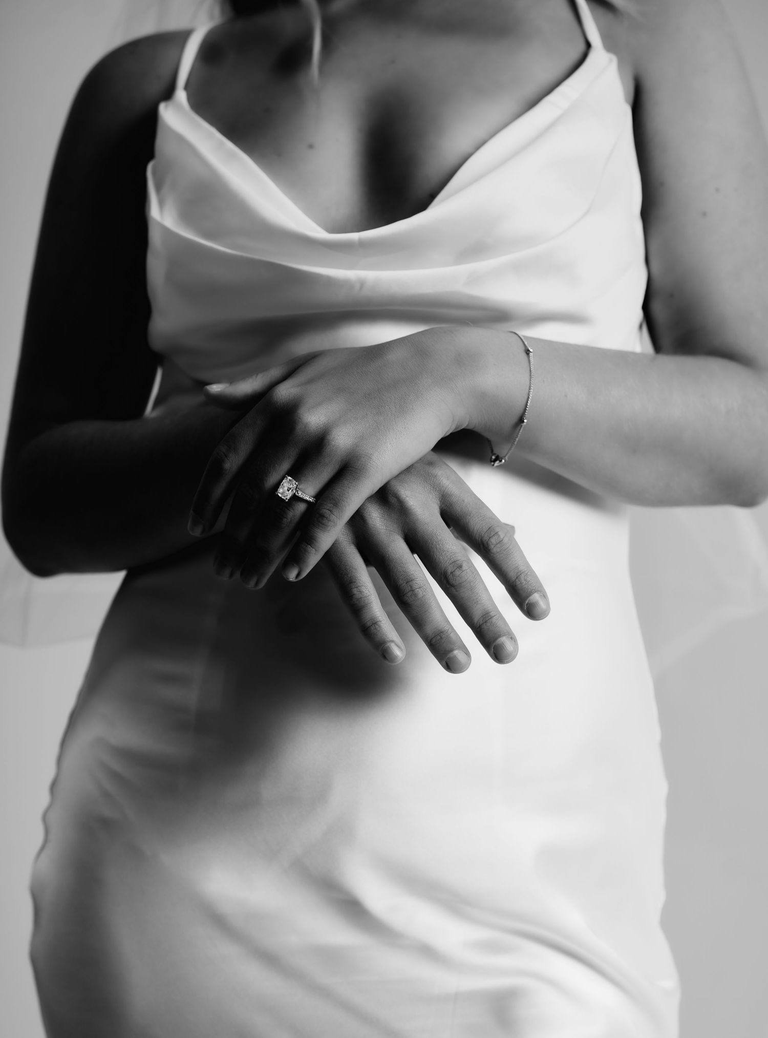 Black and white photo of a bride in a white slip dress, hands crossed showing an engagement ring and bracelet.