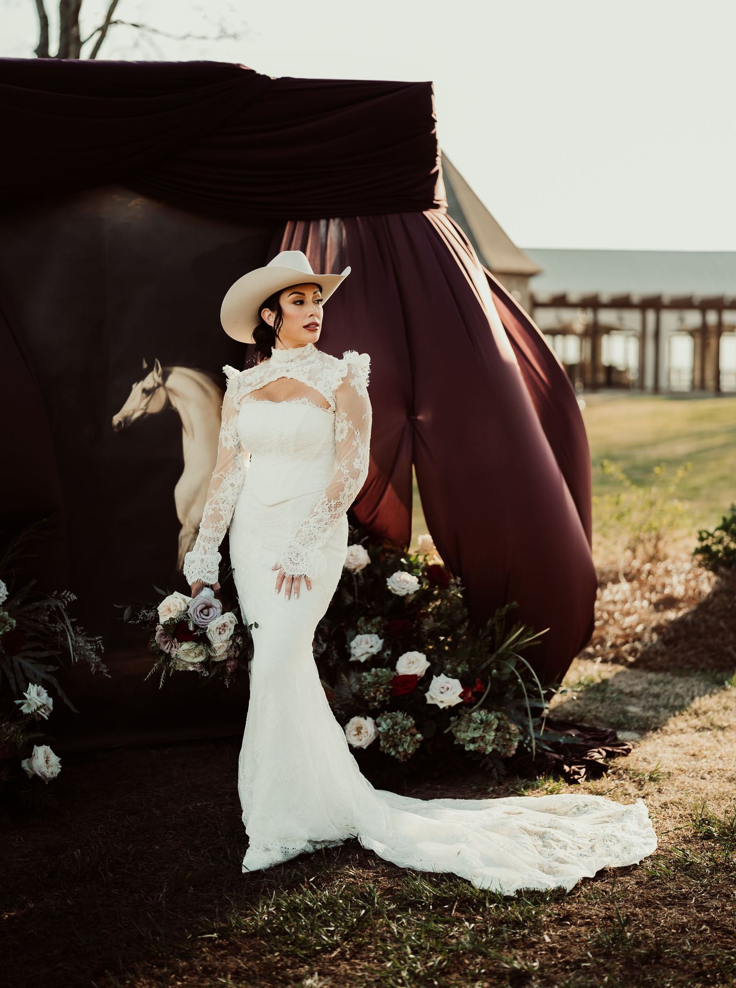 Bride in white lace gown and cowboy hat poses outdoors near floral arrangement and dark draped backdrop.