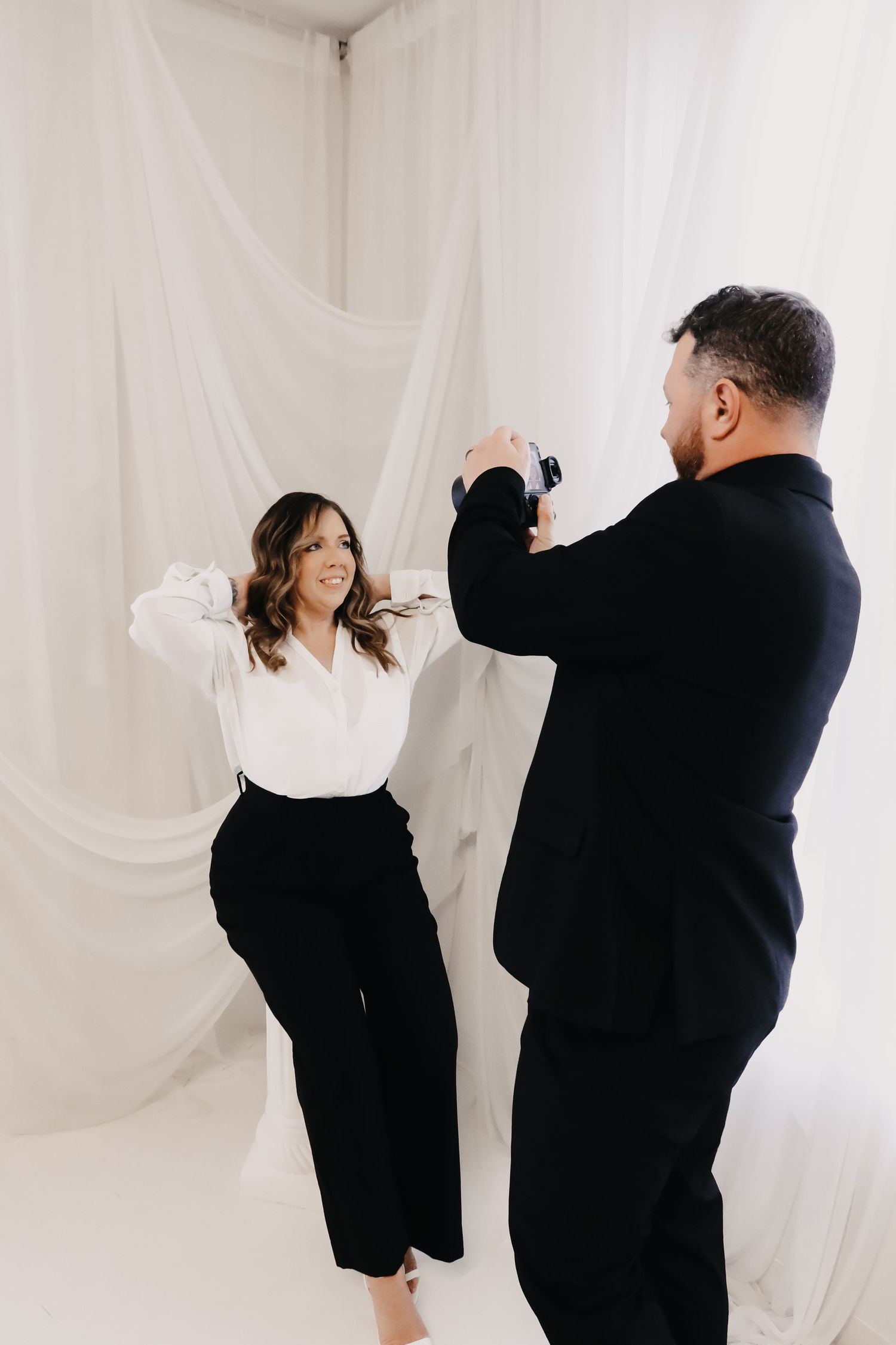 A photographer in a black suit captures a smiling woman in white blouse and black pants posing in a white draped studio.