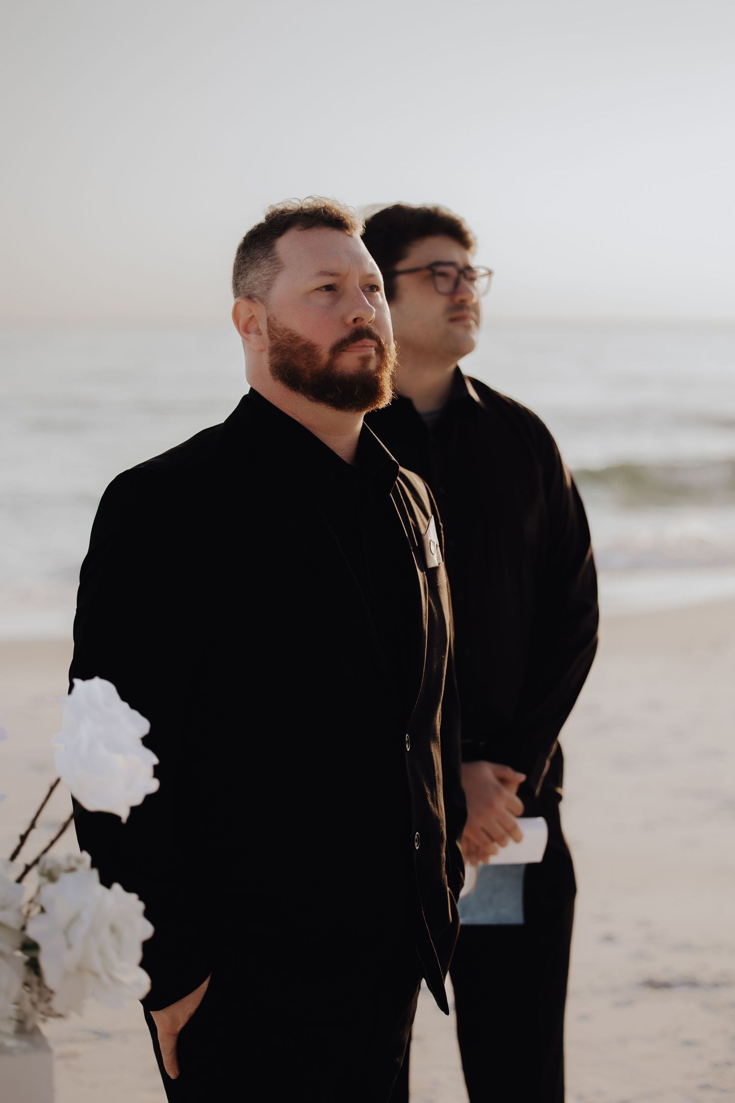 Two people in black clothing walking together on a beach with waves in the background.