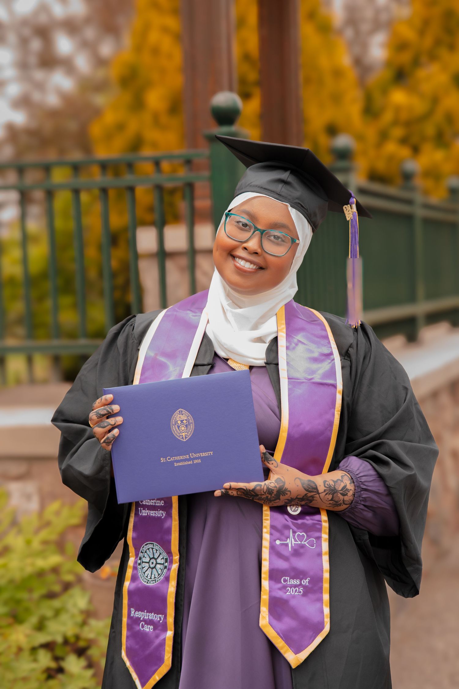 A graduate in academic regalia holding a diploma in front of fall foliage.